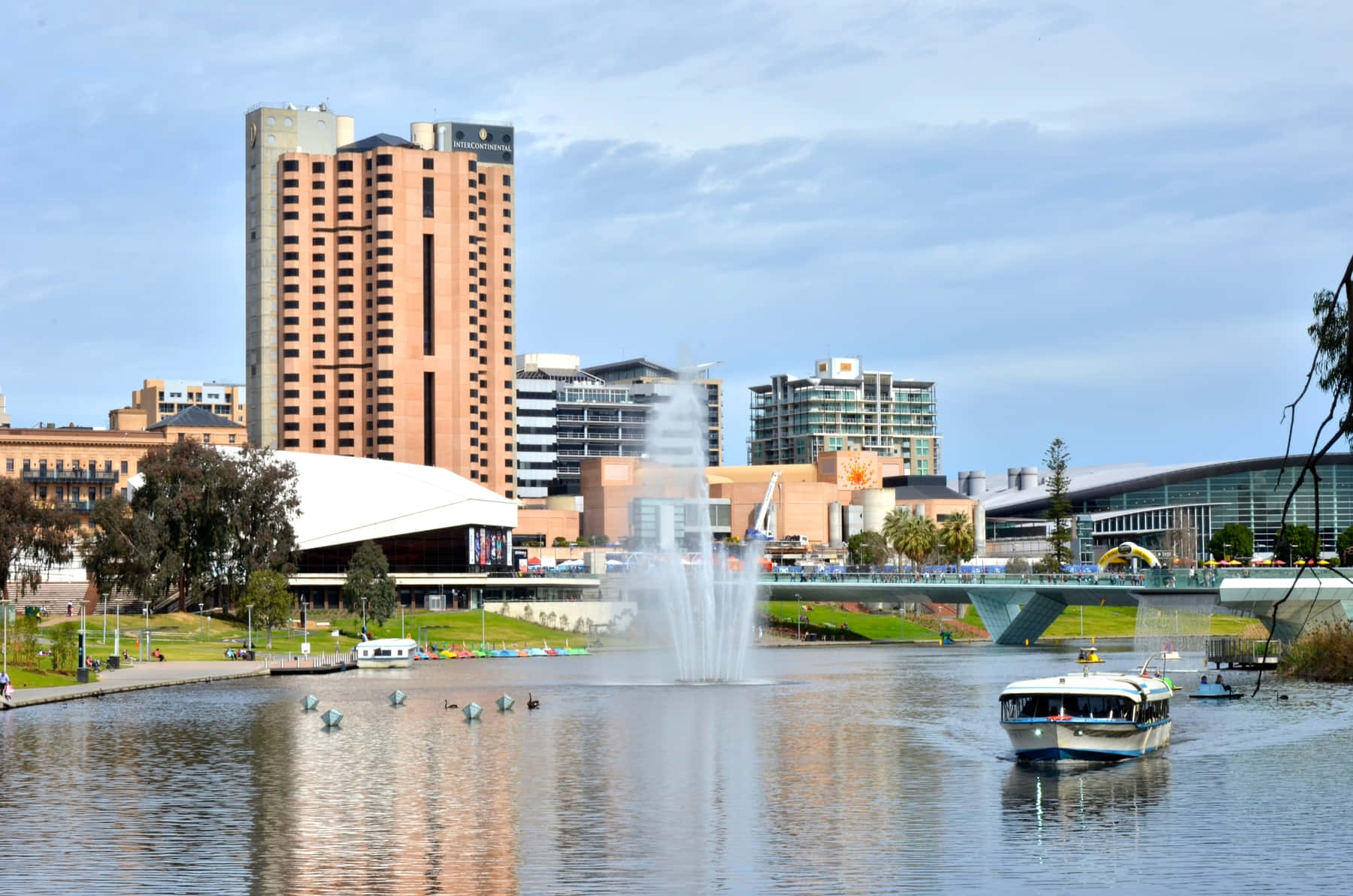 Adelaide 桌布 壁紙 — 一個充滿活力的城市海濱場景，有一座高磚飯店、中央噴泉和船隻在部分多雲的天空下平靜的水面上滑行。
