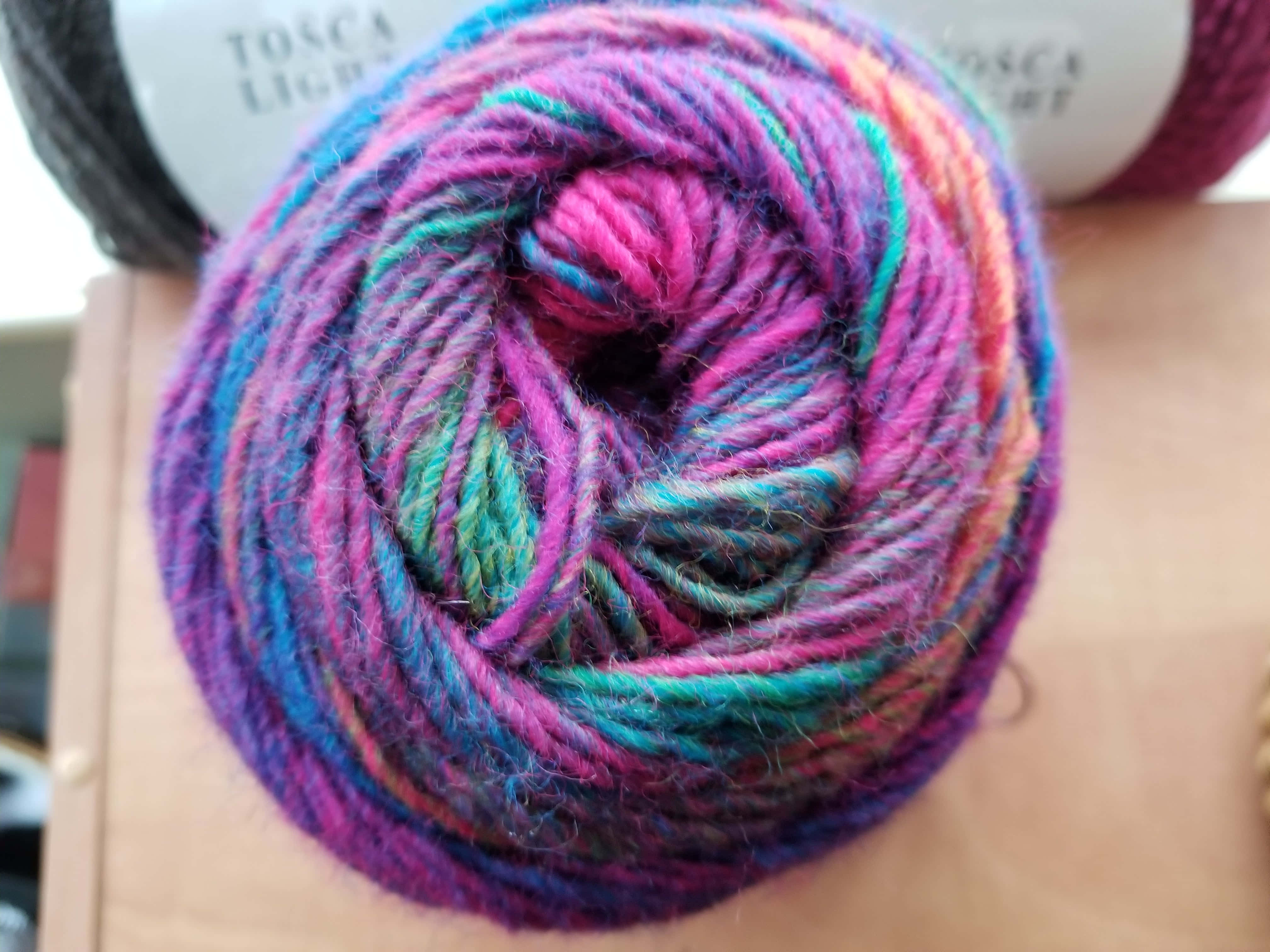 Knitting 桌布 壁紙 — 一個充滿活力的多色毛線球，帶有粉紅色、藍色和藍綠色色調，放在木質表面上，背景有其他毛線球。