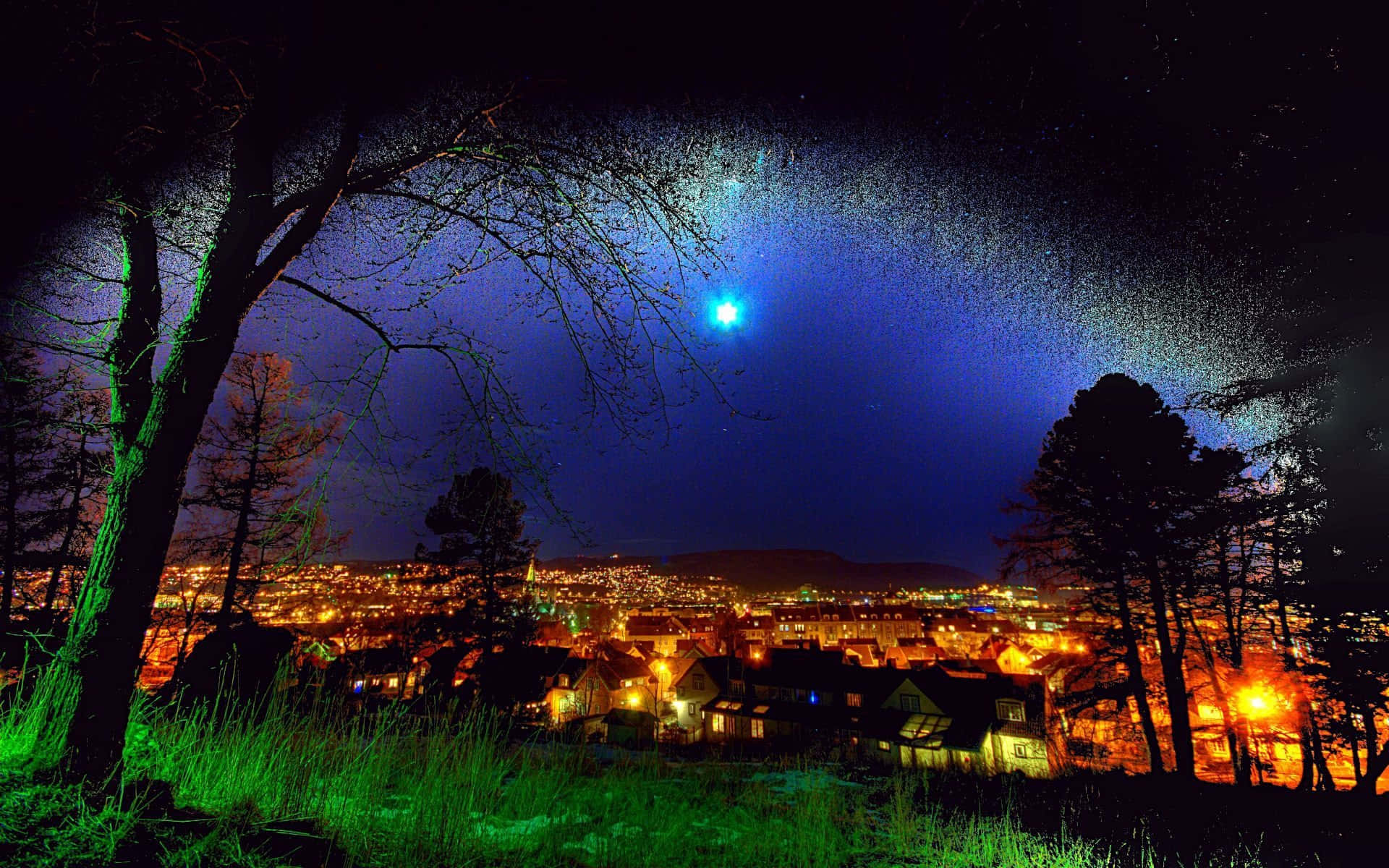 Blue Sky 桌布 壁紙 — 一個充滿活力的夜景，一顆發光的藍色星星位於城鎮上方，城鎮被溫暖的黃色燈光照亮，周圍環繞著深色樹木和綠色草地。