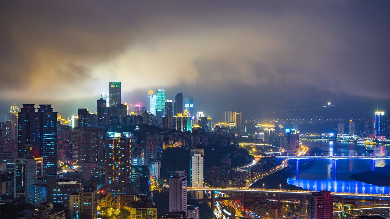 Chongqing 桌布 壁紙 — 一個充滿活力的夜景城市景觀，在戲劇性的多雲天空下，河流上有發光的摩天大樓和橋樑。