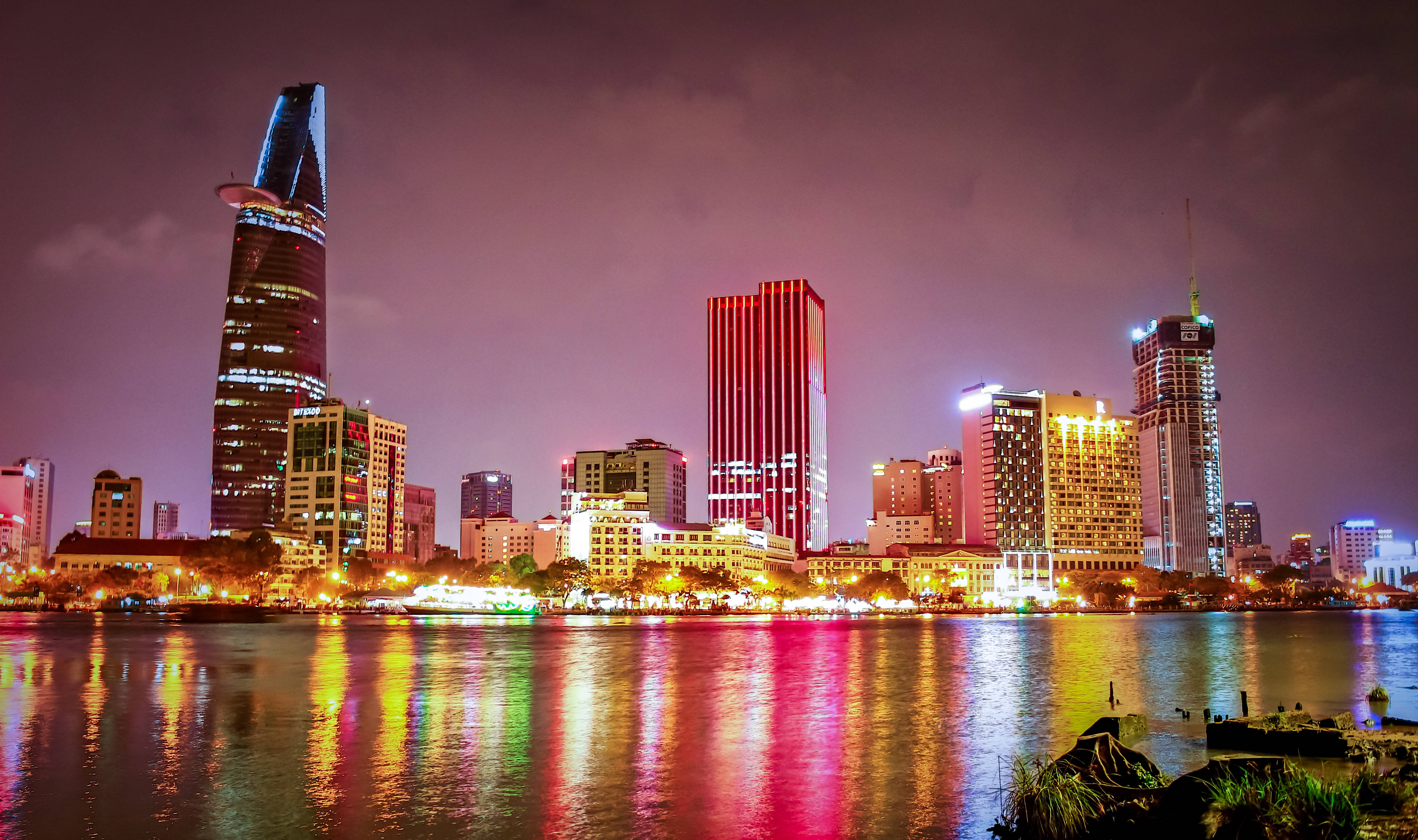 越南 桌布 壁紙 — 一個充滿活力的夜景城市景觀，在昏暗的紫色天空下，發光的摩天大樓倒映在平靜的河流中。