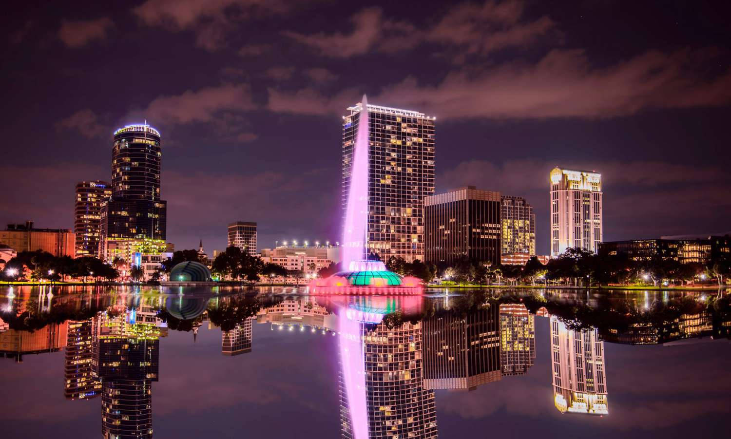 lights 桌布 壁紙 — 一個充滿活力的夜景城市景觀，在陰天之下，平靜的水面映照著燈火通明的摩天大樓和色彩繽紛的噴泉。
