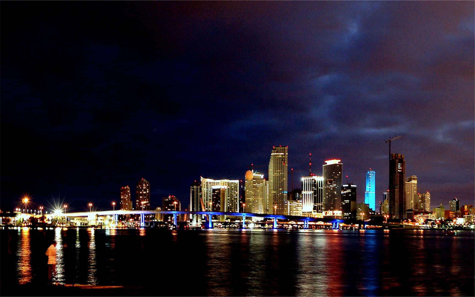 Miami 桌布 壁紙 — 一個充滿活力的夜景城市景觀，在陰天之下，漆黑的湖面倒映著燈火通明的摩天大樓和一座發光的橋樑。