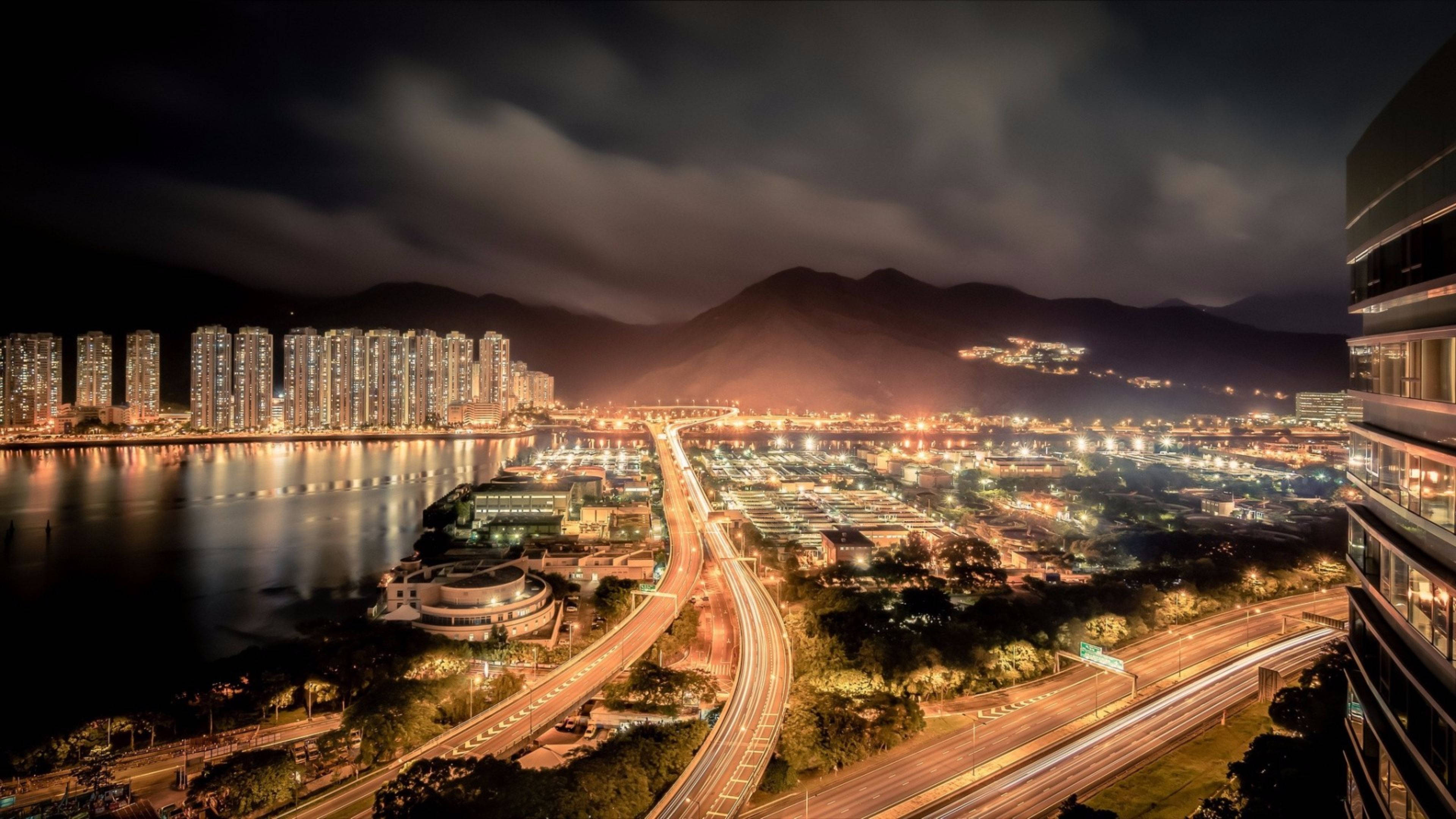 Trending 桌布 壁紙 — 一個充滿活力的夜景城市景觀，有發光的摩天大樓、照明的高速公路和遠處的山脈，天空多雲。