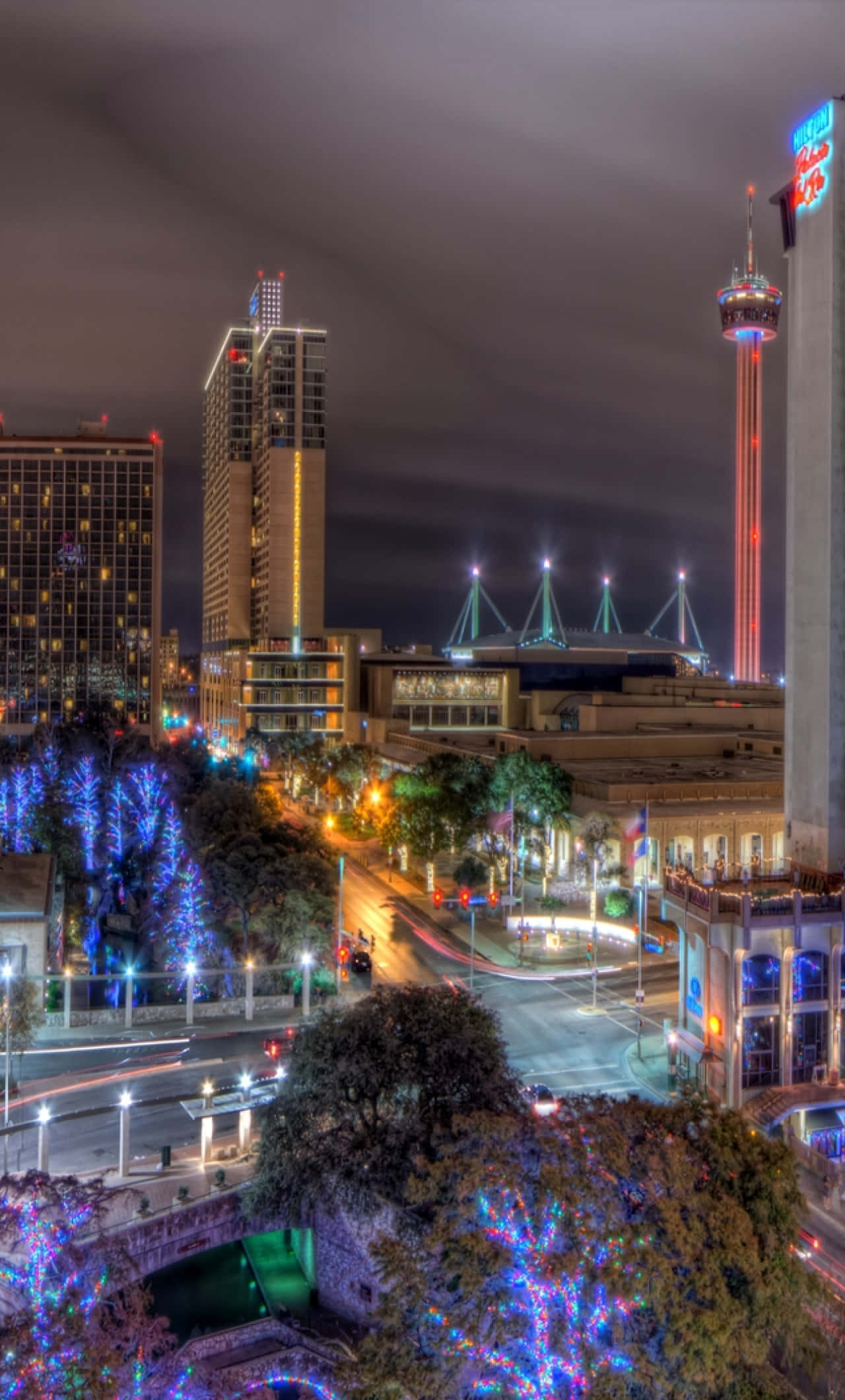 San Antonio 桌布 壁紙 — 一個充滿活力的夜景城市景觀，特色是燈火通明的摩天大樓和樹上節日藍色燈光，在黑暗的天空下。