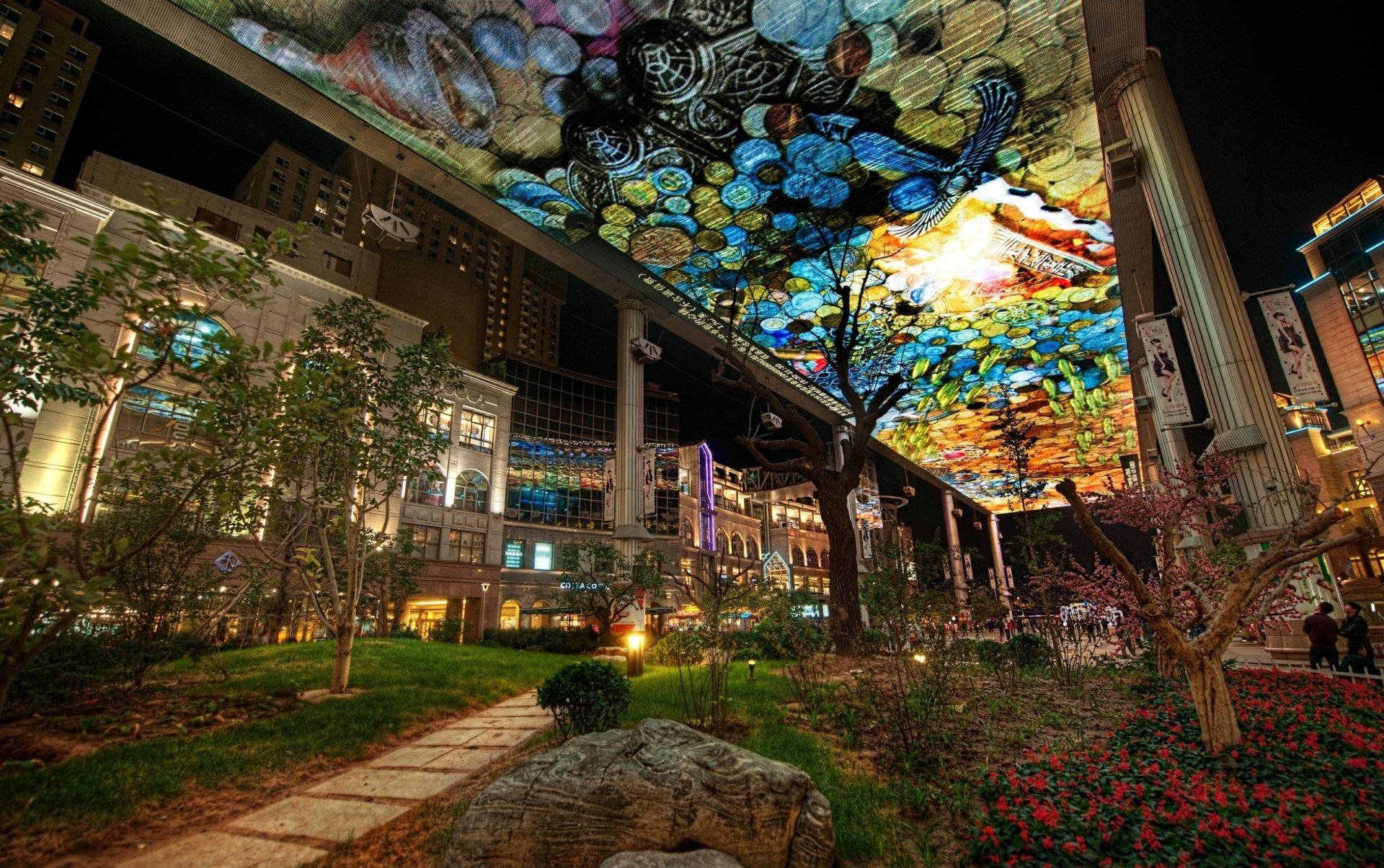 Beijing 桌布 壁紙 — 一個充滿活力的夜間城市廣場，在花園小徑上有一個巨大的彩色數位天篷投影，小徑上有樹木和花朵。