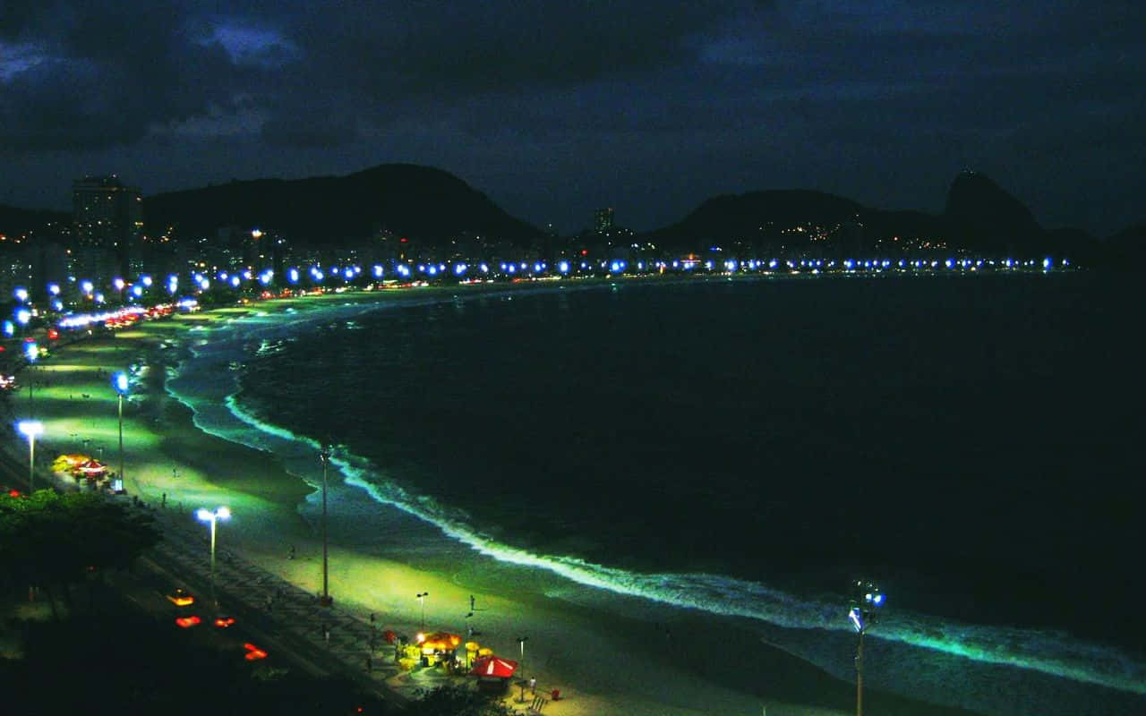 巴西 桌布 壁紙 — 一個充滿活力的夜間海灘場景，有發光的藍綠色海浪、燈火通明的木板路，以及遠處城市燈光映襯著黑暗的山丘。