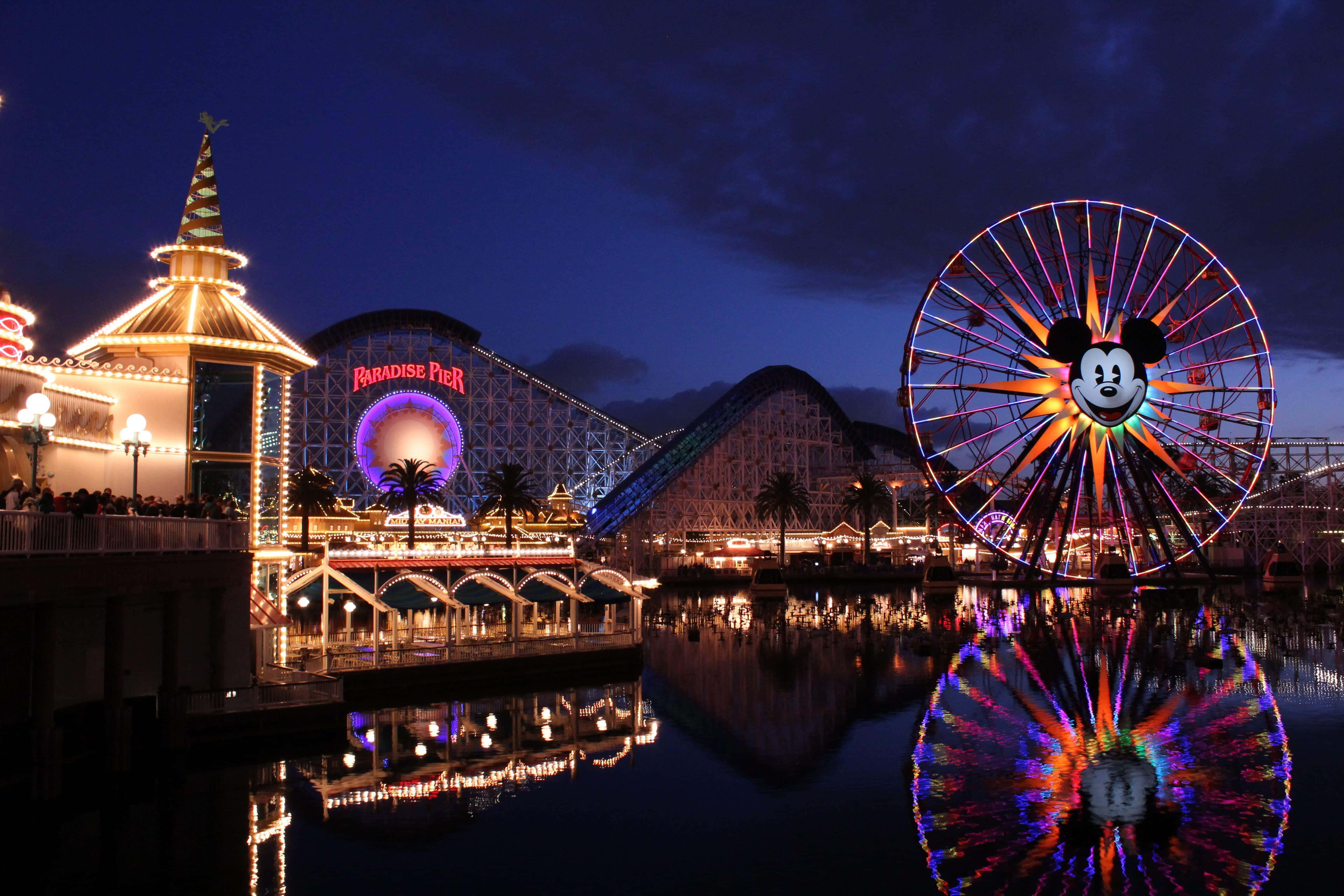 Disneyland 桌布 壁紙 — 一個充滿活力的夜間遊樂園，發光的米奇老鼠摩天輪和雲霄飛車倒映在平靜的水面上。