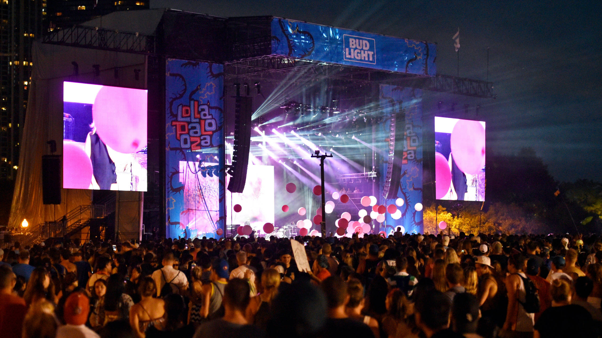 Lollapalooza 桌布 壁紙 — 一個充滿活力的夜間音樂會舞台，沐浴在紫色和粉紅色的燈光中，下方擠滿了人群。
