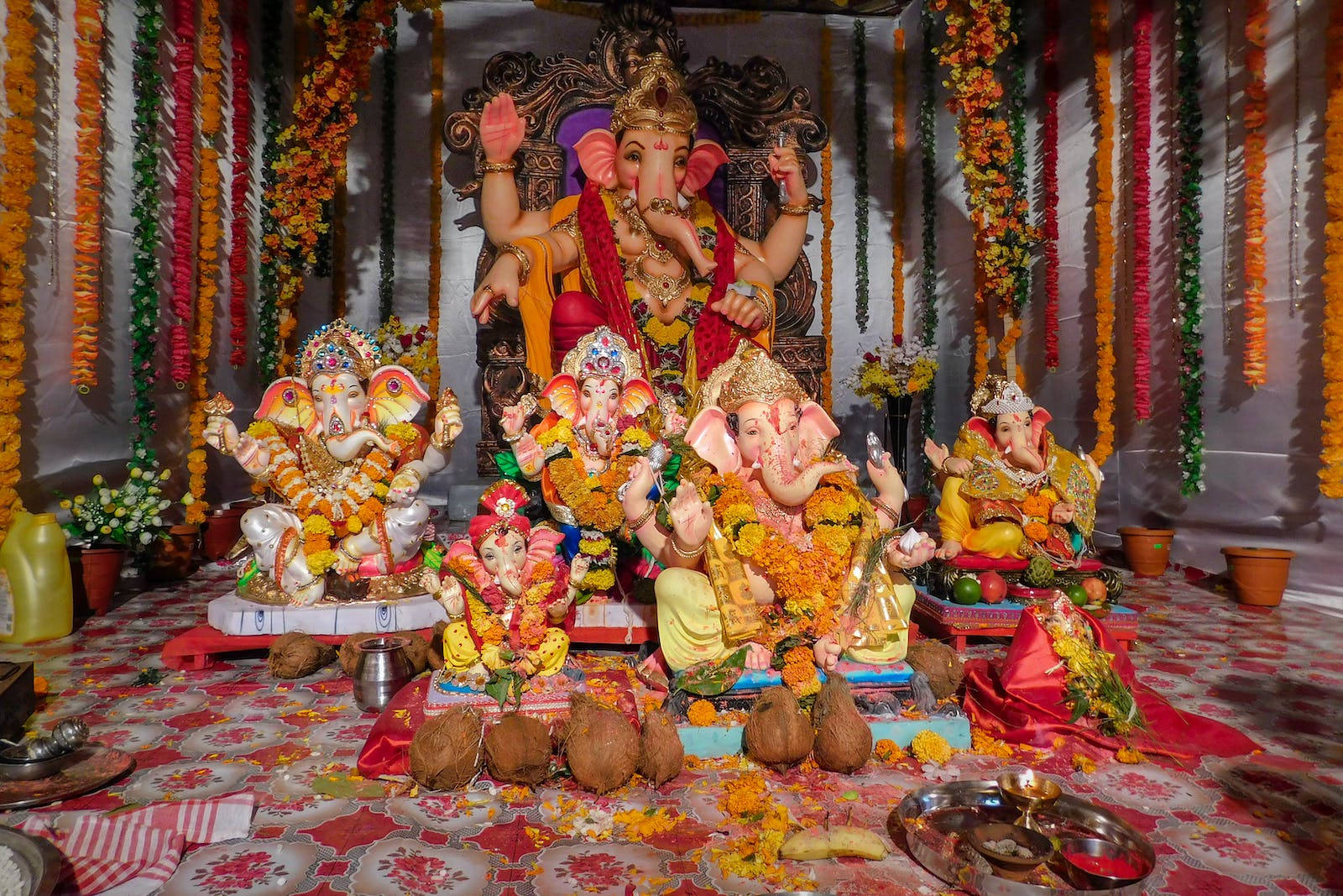 Vinayaka 桌布 壁紙 — 一個充滿活力的室內神龕展示多個色彩繽紛的象神雕像，裝飾著萬壽菊和供品，懸掛在節日花環下。