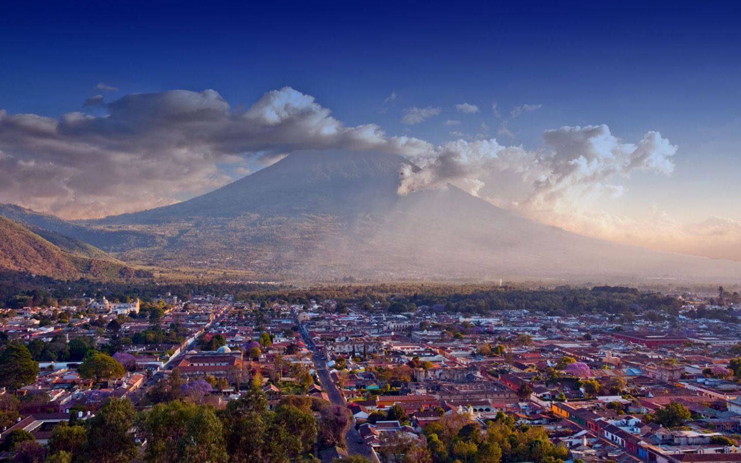 Guatemala 桌布 壁紙 — 一個充滿活力的小鎮，在柔和的雲彩和戲劇性的日出天空下，坐落在一座雄偉的火山腳下。