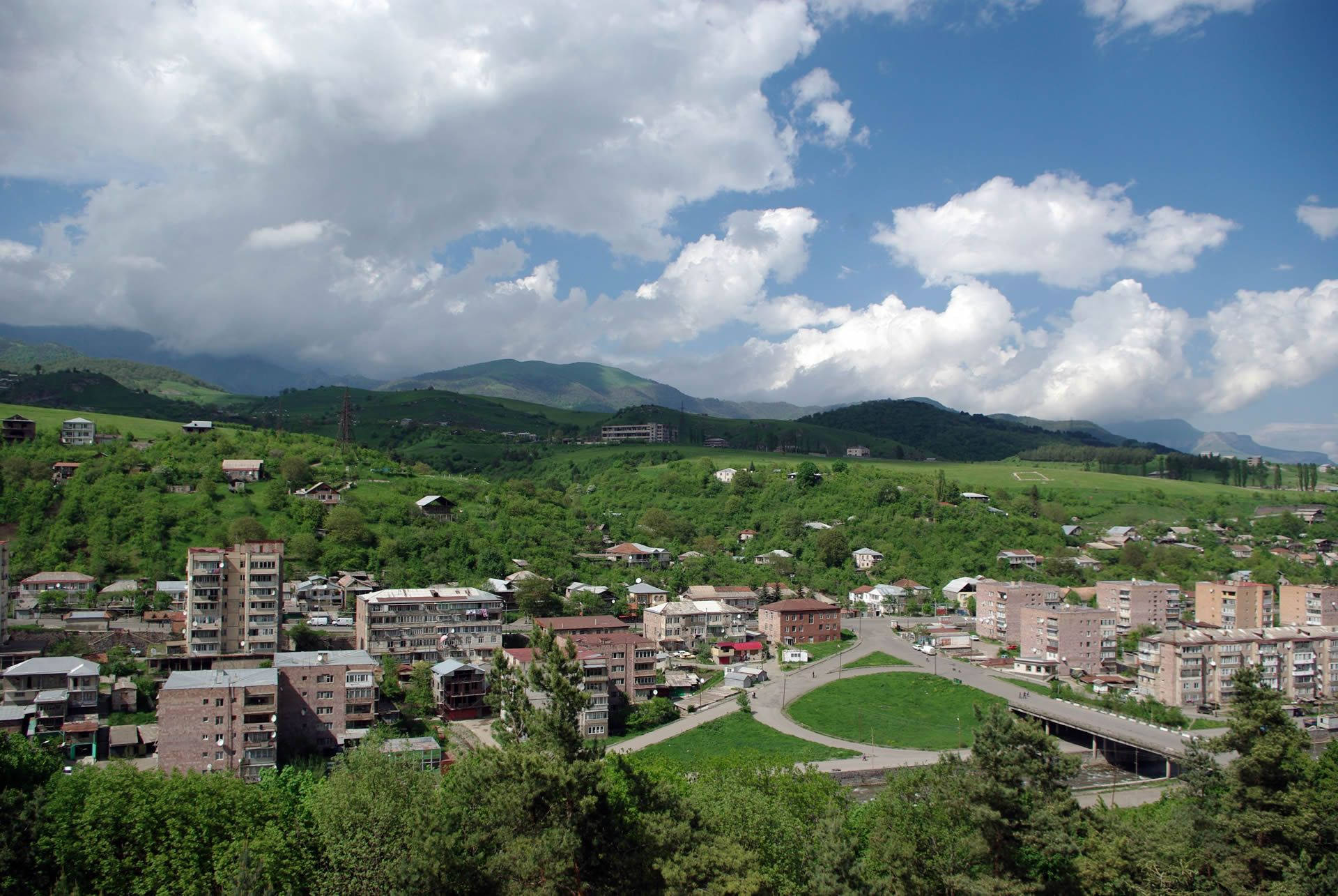 Armenia 桌布 壁紙 — 一個充滿活力的小鎮，坐落在鬱鬱蔥蔥的綠色山丘中，上方是明亮的藍天和蓬鬆的白雲。