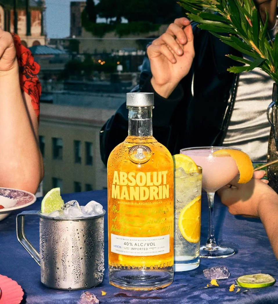 bottle 桌布 壁紙 — 一個充滿活力的屋頂雞尾酒場景，包含 Absolut Mandrin 酒瓶和帶有柑橘裝飾的飲品，放在藍色桌布上。