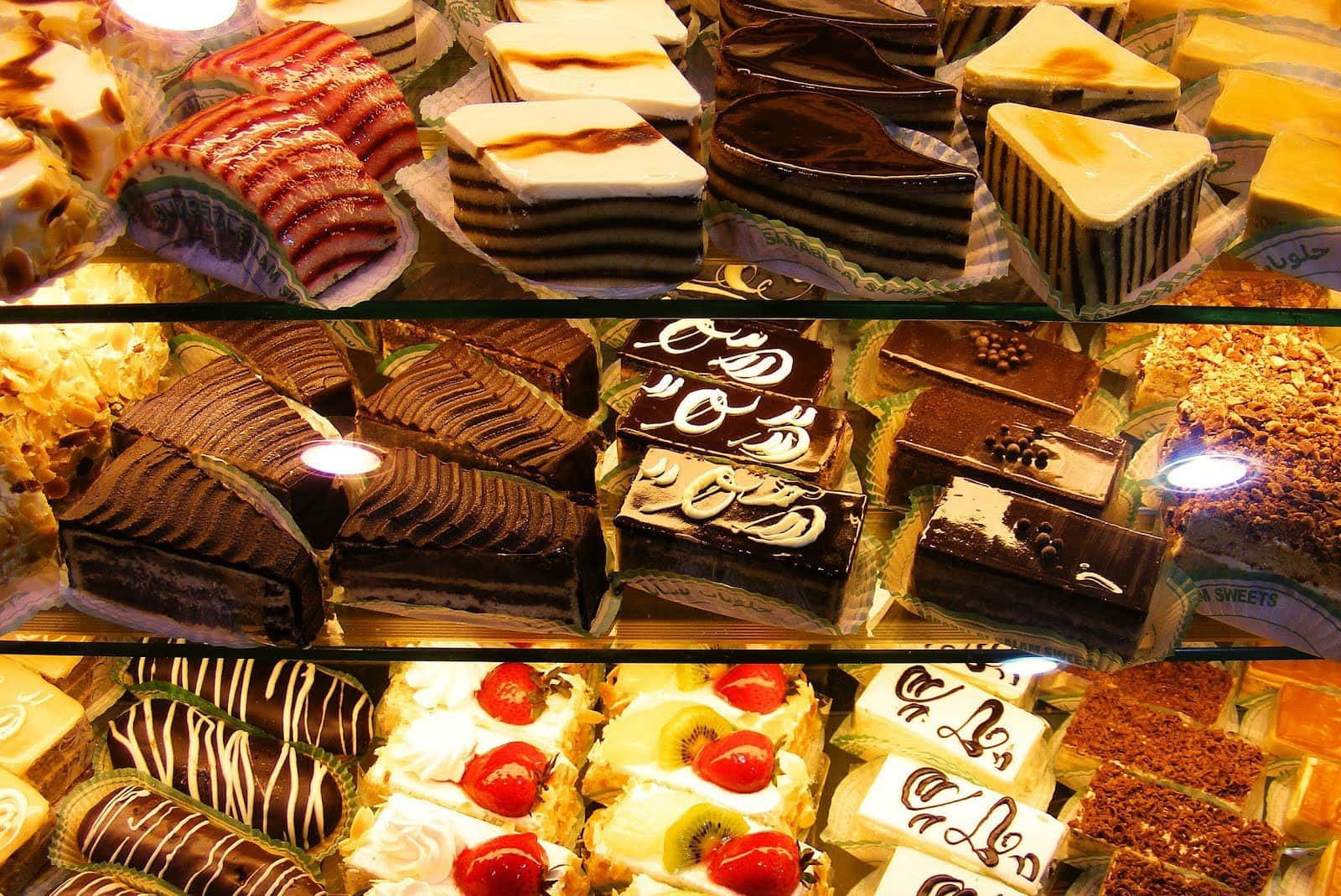 Pastry 桌布 壁紙 — 一個充滿活力的展示櫃，裡面擺滿了各式各樣的夾層蛋糕和糕點，在溫暖的燈光下閃閃發光。