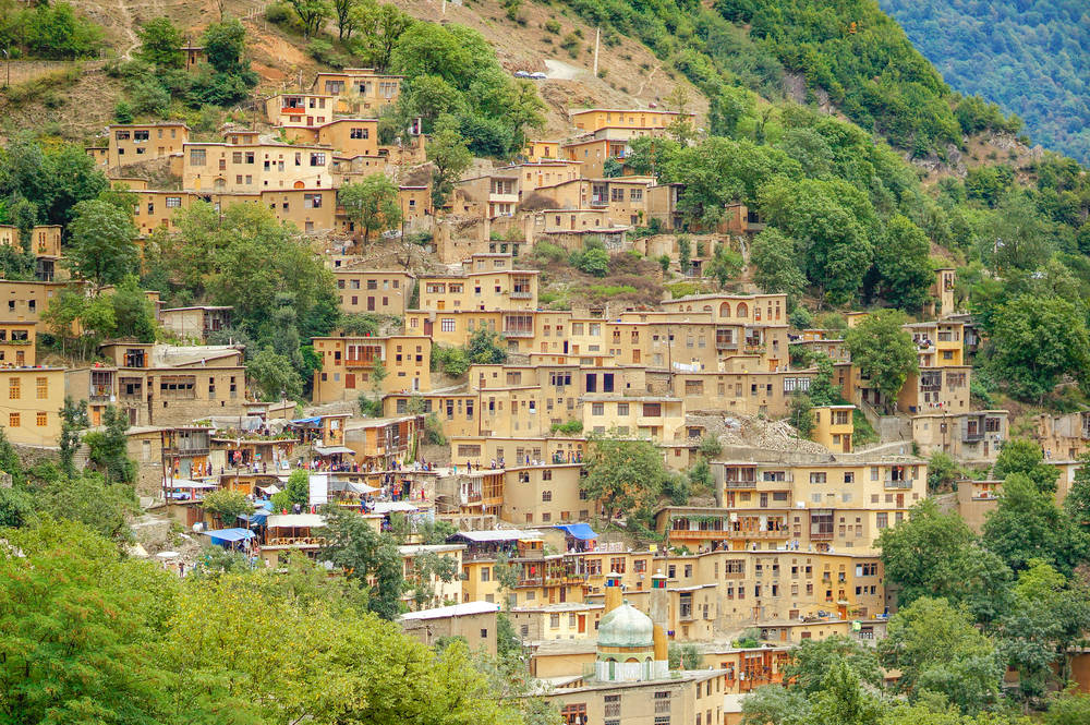 Iran 桌布 壁紙 — 一個充滿活力的山坡村莊，赤陶建築坐落在茂密的綠樹之間，沐浴在明亮的日光下。