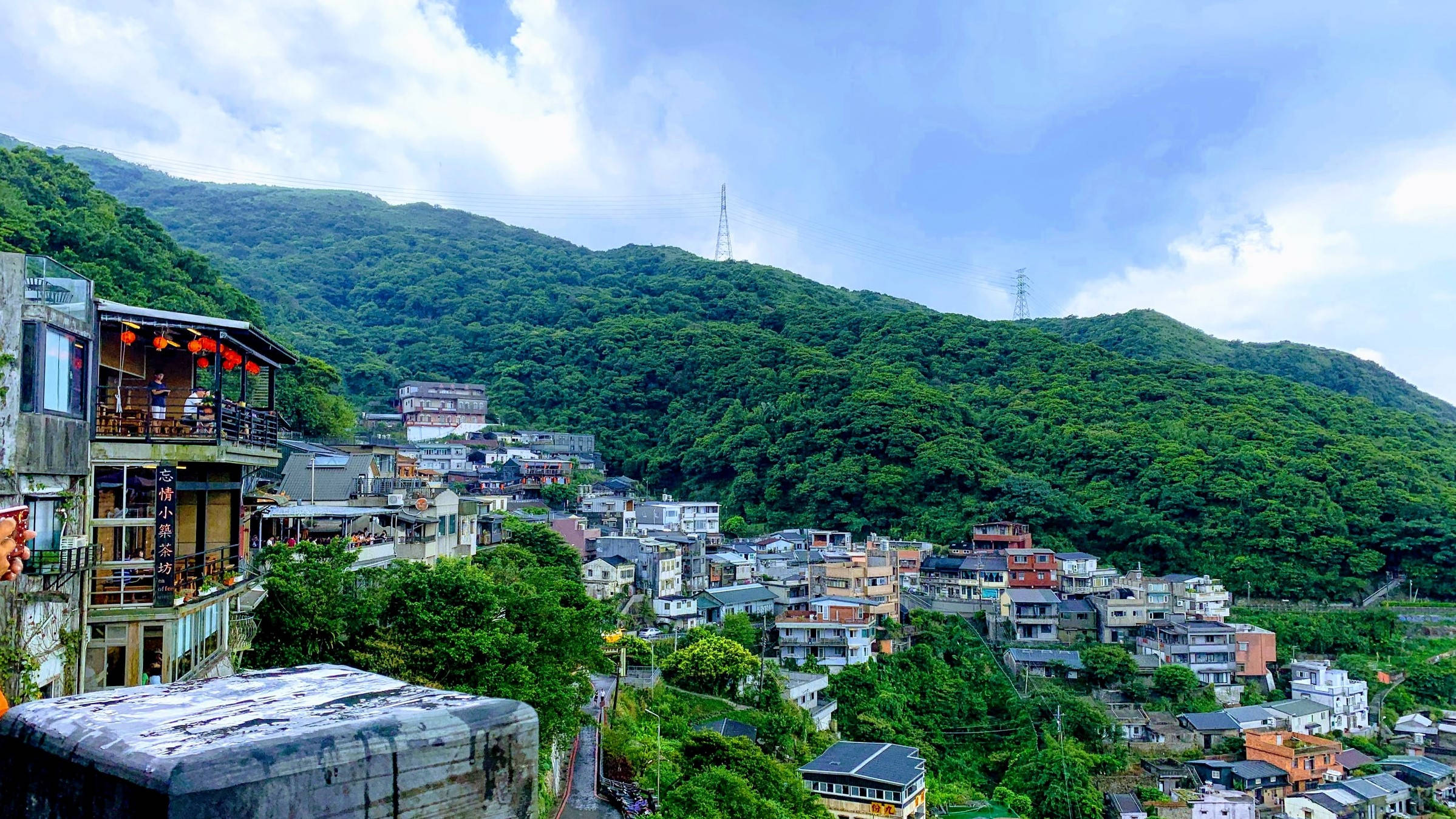 台北 桌布 壁紙 — 一個充滿活力的山坡村莊依附在鬱鬱蔥蔥的綠色山脈上，天空湛藍，雲朵點綴。