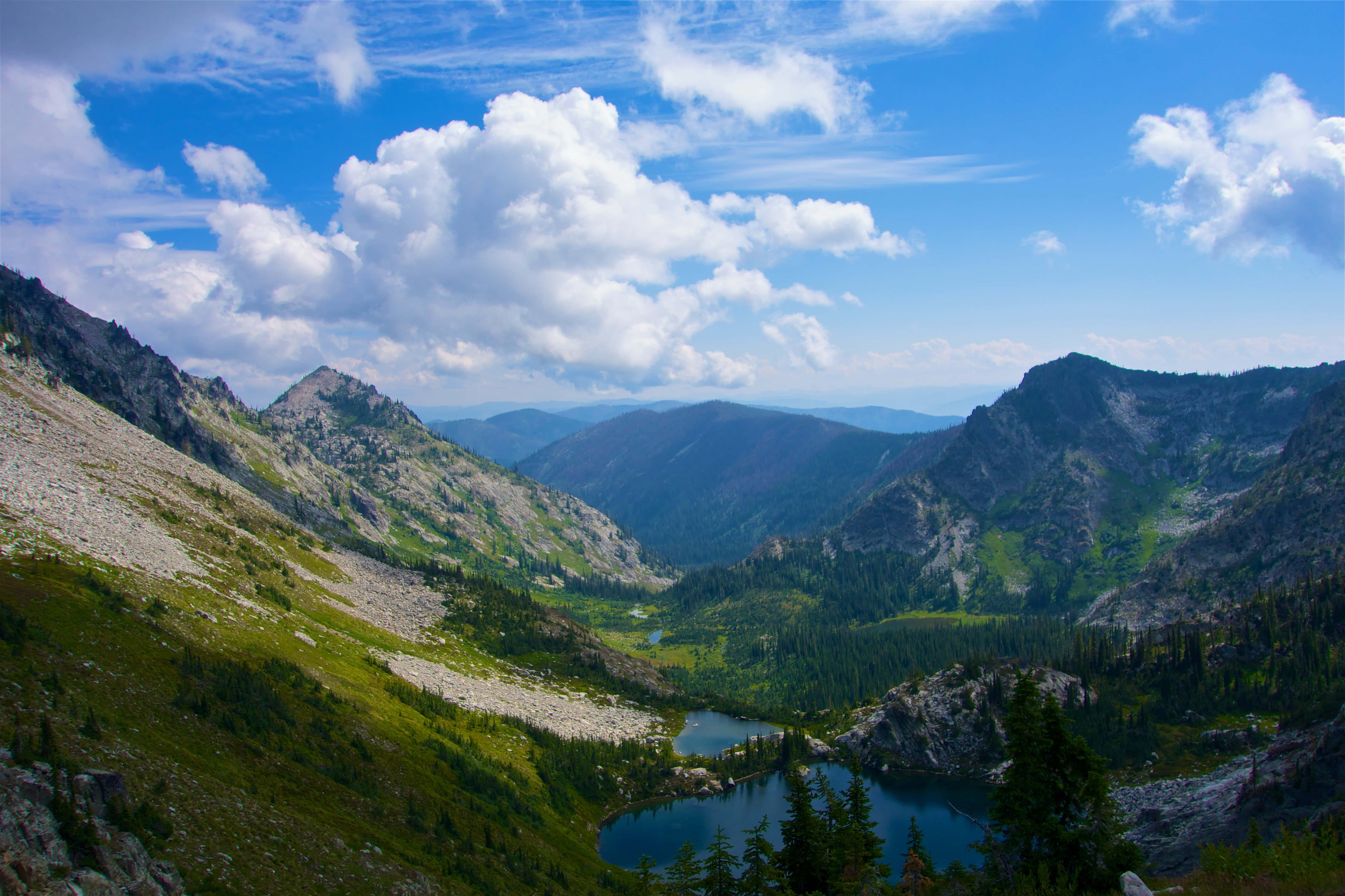 Beautiful View 桌布 壁紙 — 一個充滿活力的山谷，擁有深藍色的湖泊，坐落在岩石山峰和茂密的綠色森林之間，在明亮的藍天下有蓬鬆的白雲。