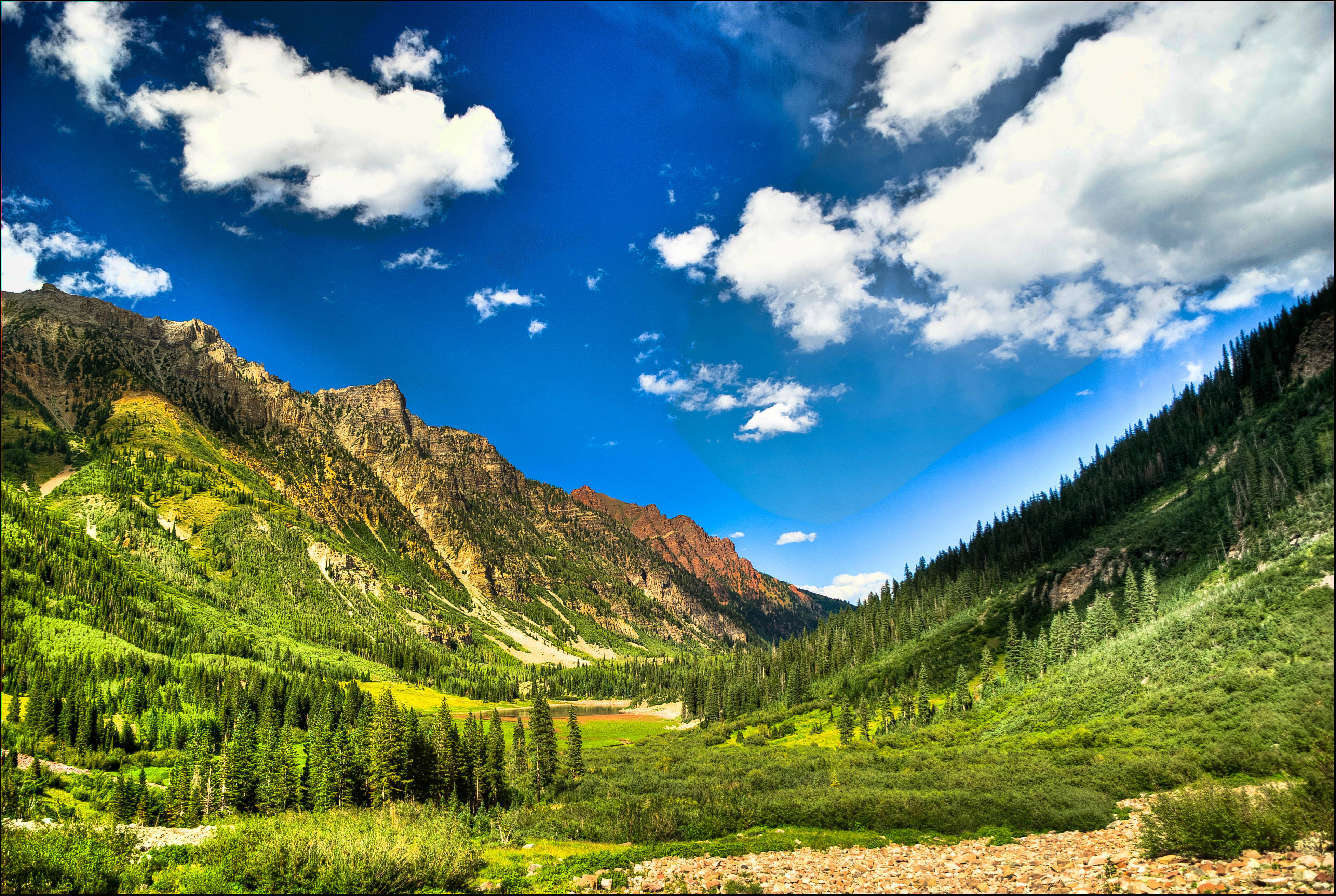 Colorado 桌布 壁紙 — 一個充滿活力的山谷，擁有茂密的綠色森林和岩石山峰，在明亮的藍天下有蓬鬆的白雲。