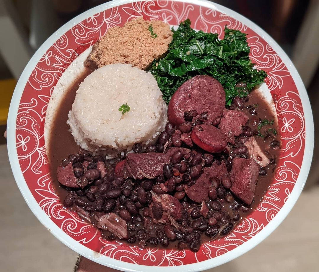 yummy 桌布 壁紙 — 一個充滿活力的巴西黑豆燉肉碗，配有黑豆、煙燻肉、白飯和青菜，放在紅色圖案盤子上。