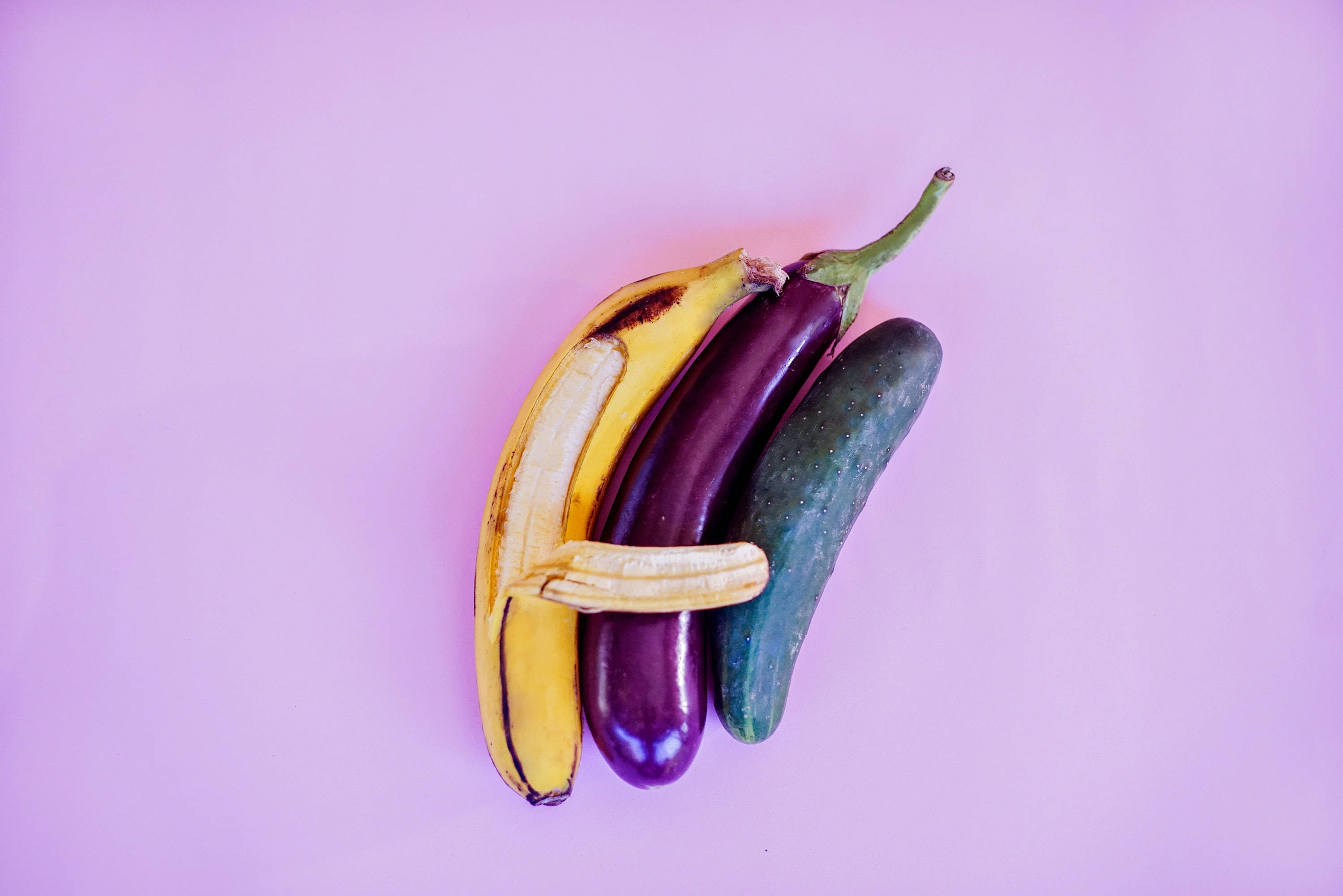 Fruits And Vegetables 桌布 壁紙 — 一個充滿活力的平鋪佈局，將香蕉、茄子和黃瓜排列在柔和的紫色背景上。