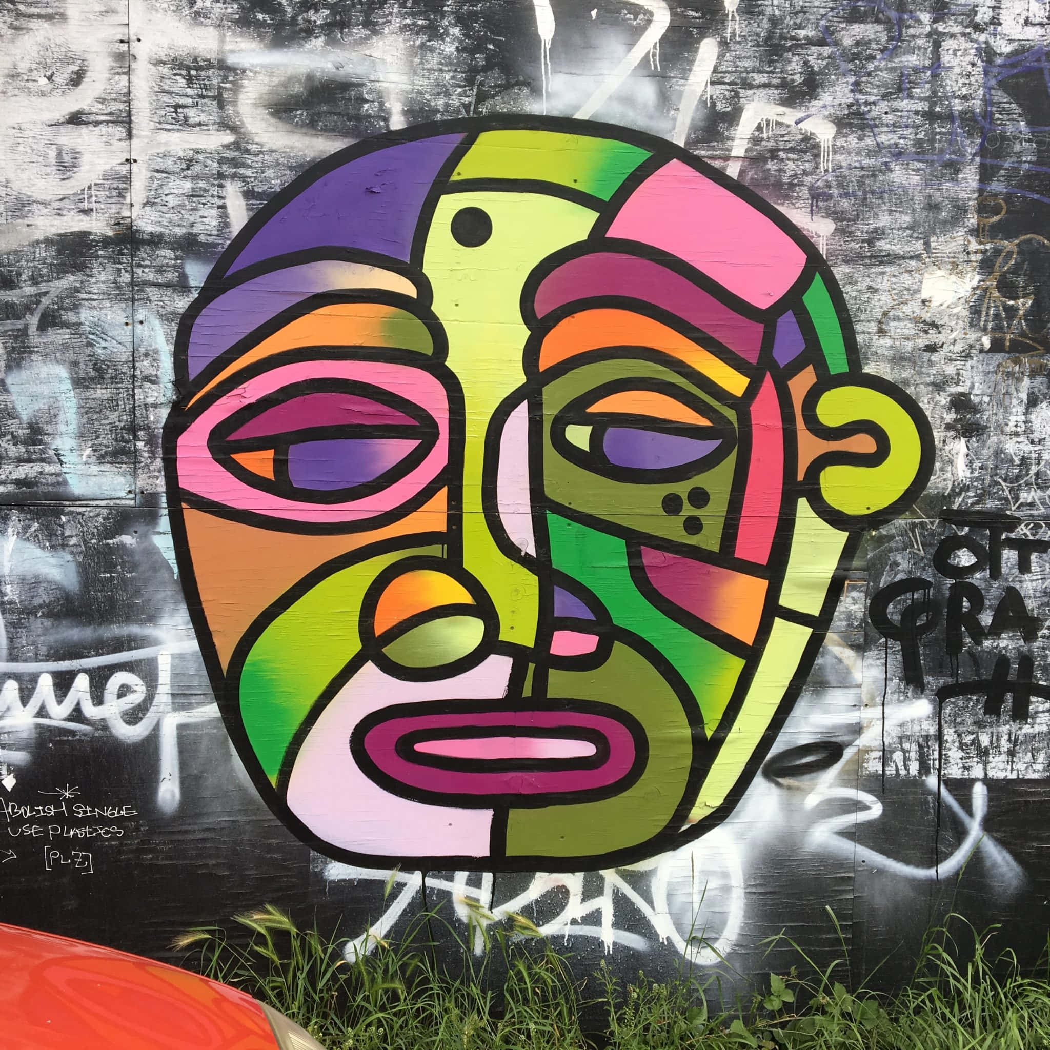 Graffiti Art 桌布 壁紙 — 一個充滿活力的幾何塗鴉面孔，帶有鮮豔色彩和黑色輪廓，畫在被草叢包圍的風化城市牆上。