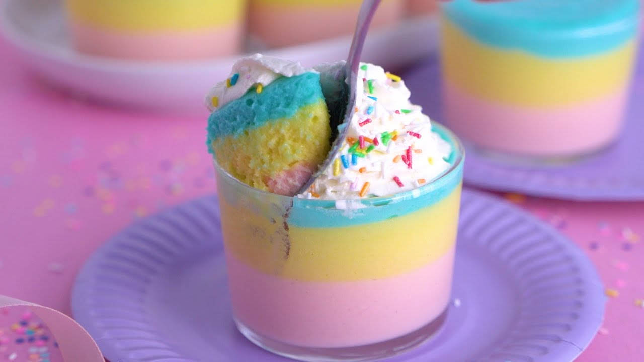 Marshmallow 桌布 壁紙 — 一個充滿活力的彩虹分層杯子蛋糕放在玻璃杯中，上面有鮮奶油和彩色糖粒，放在紫色盤子上。