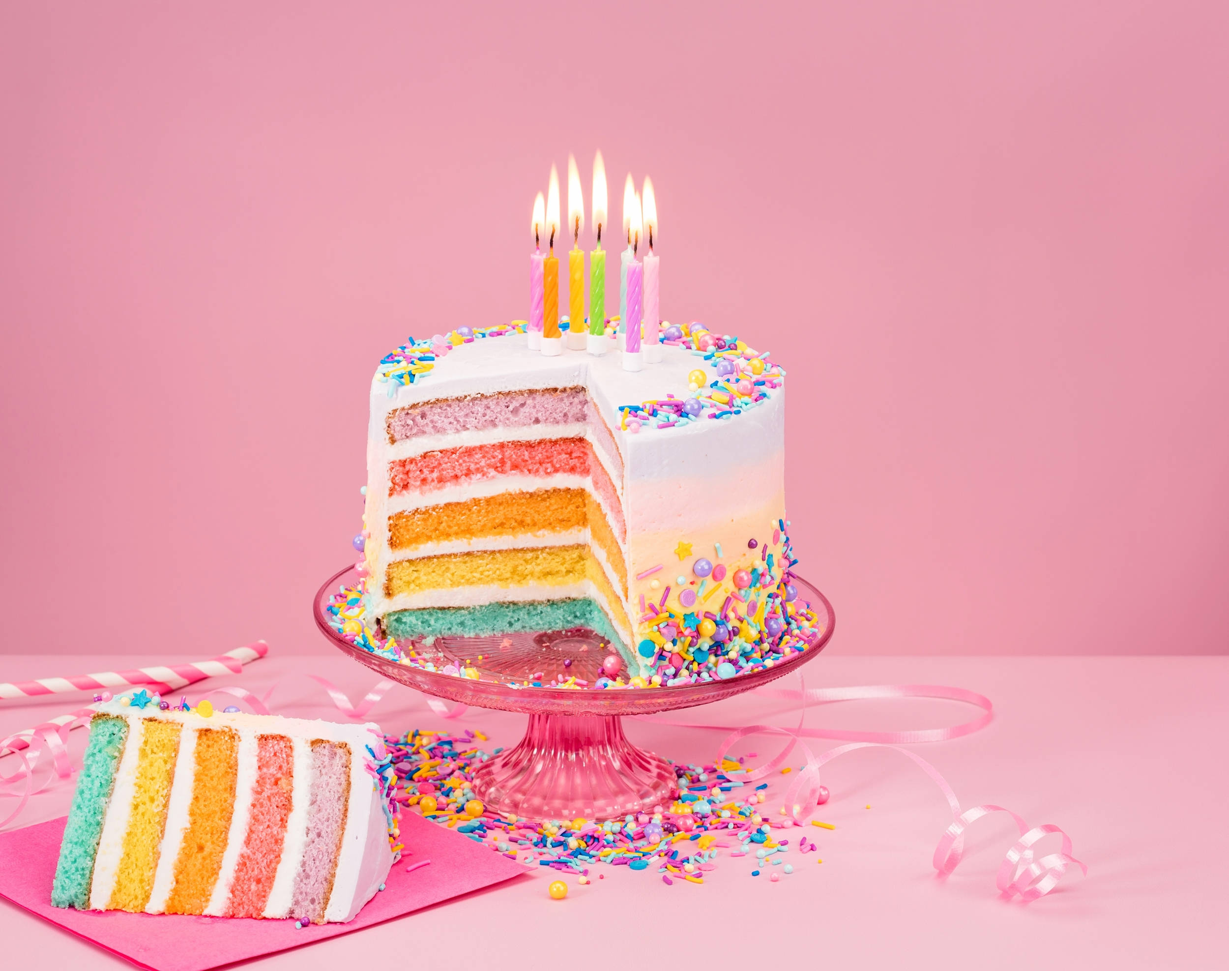 Cake 桌布 壁紙 — 一個充滿活力的彩虹分層生日蛋糕，上面有燃燒的蠟燭和彩色糖粒，放在粉紅色支架上，背景是柔和的粉紅色。
