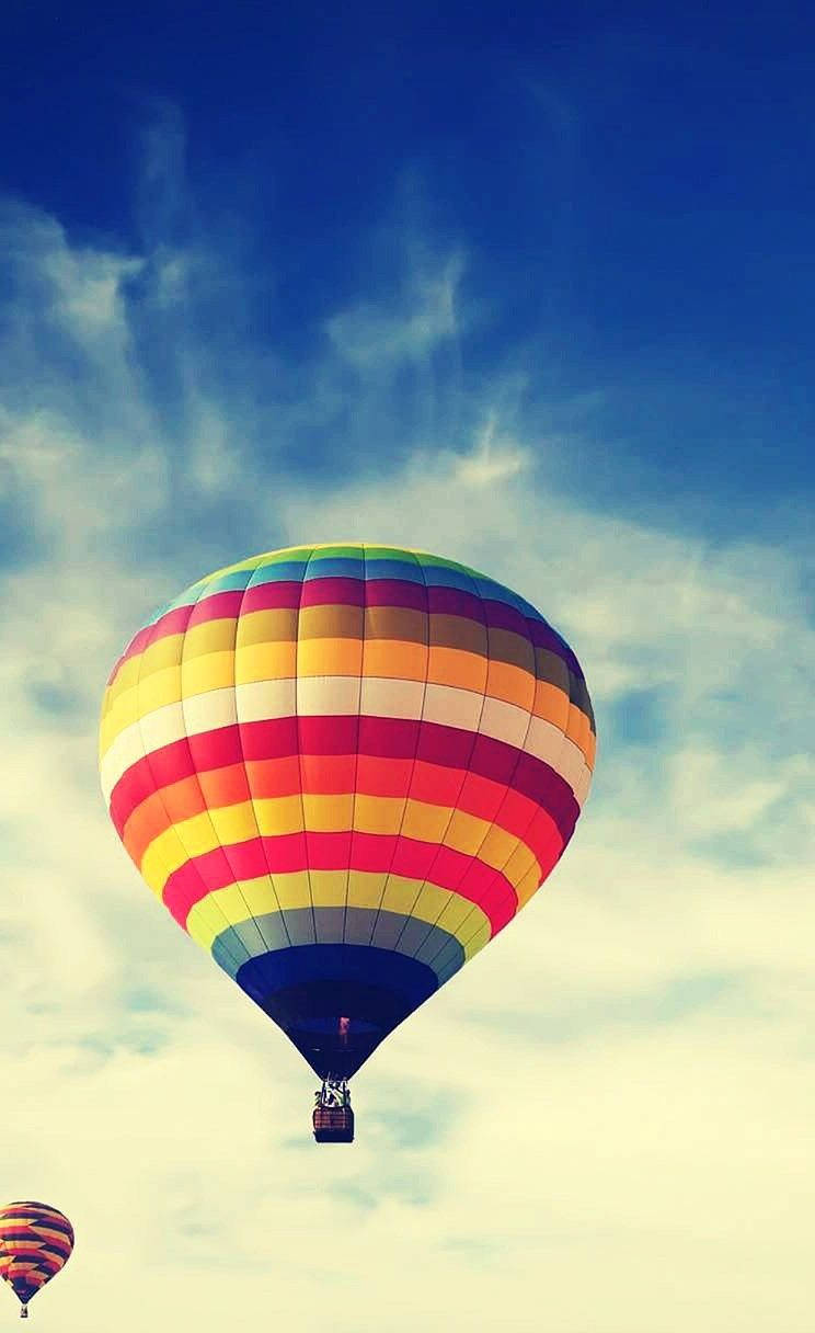 Hot Air Balloon 桌布 壁紙 — 一個充滿活力的彩虹條紋熱氣球在晴朗的藍天中飄浮，伴隨著縷縷白雲。