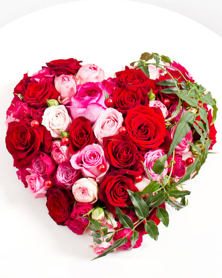 Rose Flower 桌布 壁紙 — 一個充滿活力的心形花束，由紅、粉紅、白玫瑰搭配綠葉和紅色漿果組成，置於白色表面上。
