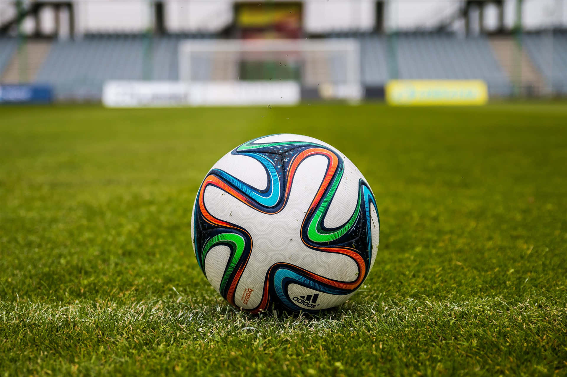 Football Field 桌布 壁紙 — 一個充滿活力的愛迪達足球放在空蕩蕩的體育館中茂密的綠草上，背景是模糊的看台和球門。
