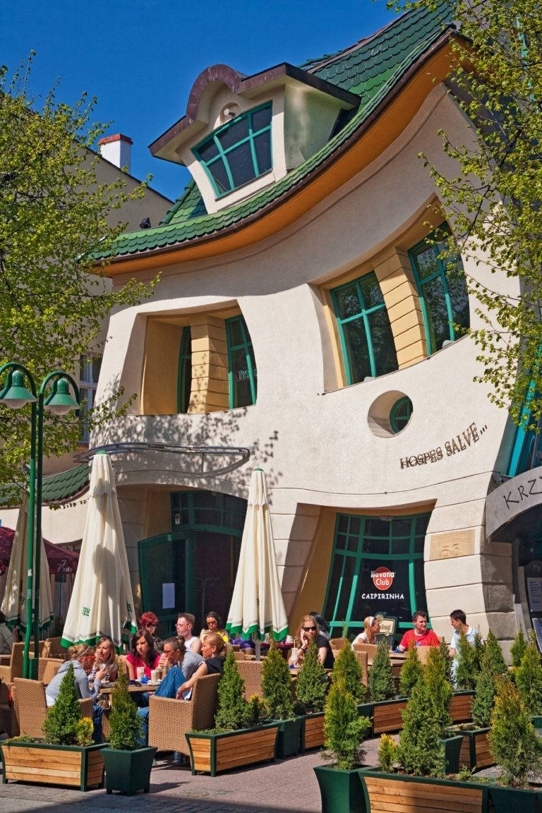 Crooked House 桌布 壁紙 — 一個充滿活力的戶外咖啡館場景，顧客在綠色遮陽傘下享受晴朗天氣，旁邊是一座帶有青綠色點綴的奇特弧形建築。