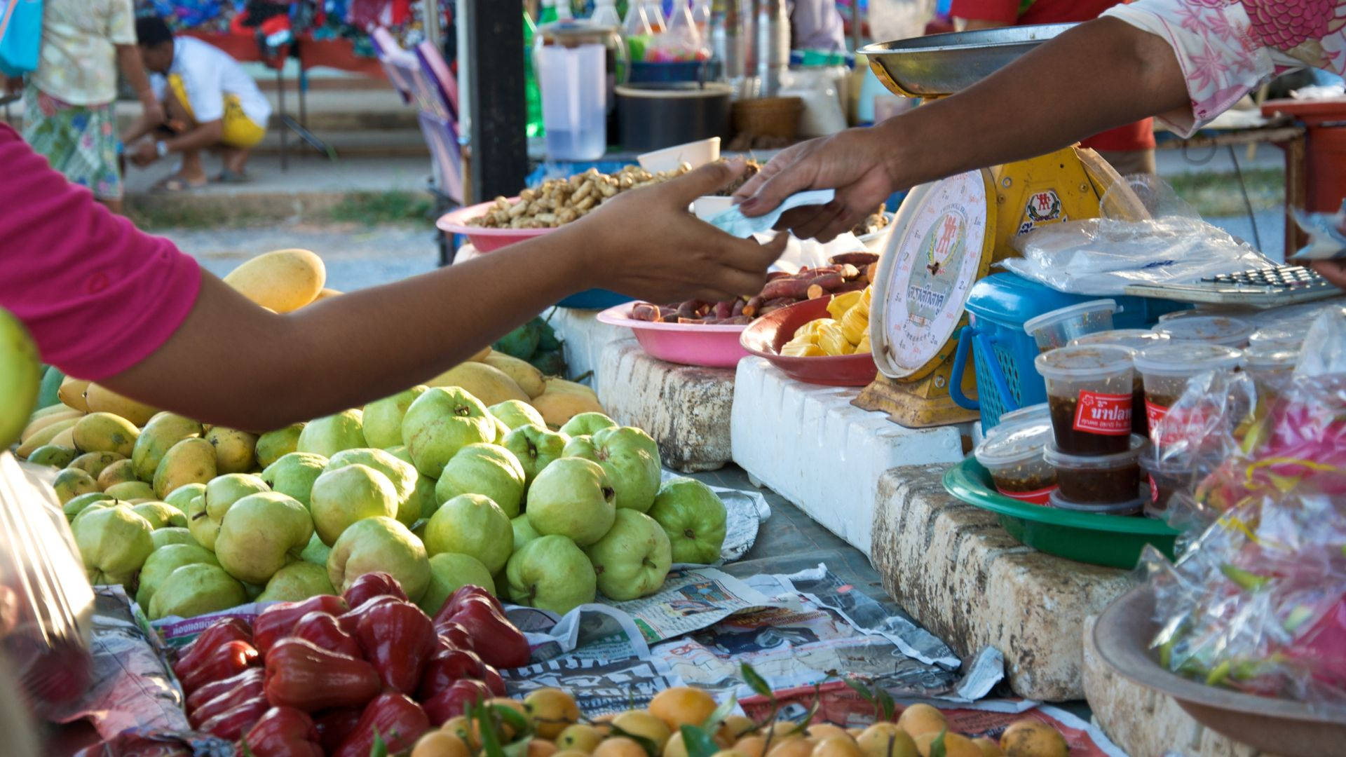 Fruits 桌布 壁紙 — 一個充滿活力的戶外市場攤位展示色彩繽紛的水果，如青色的番石榴和紅色的玫瑰蘋果，而手正在交換金錢以購買農產品。