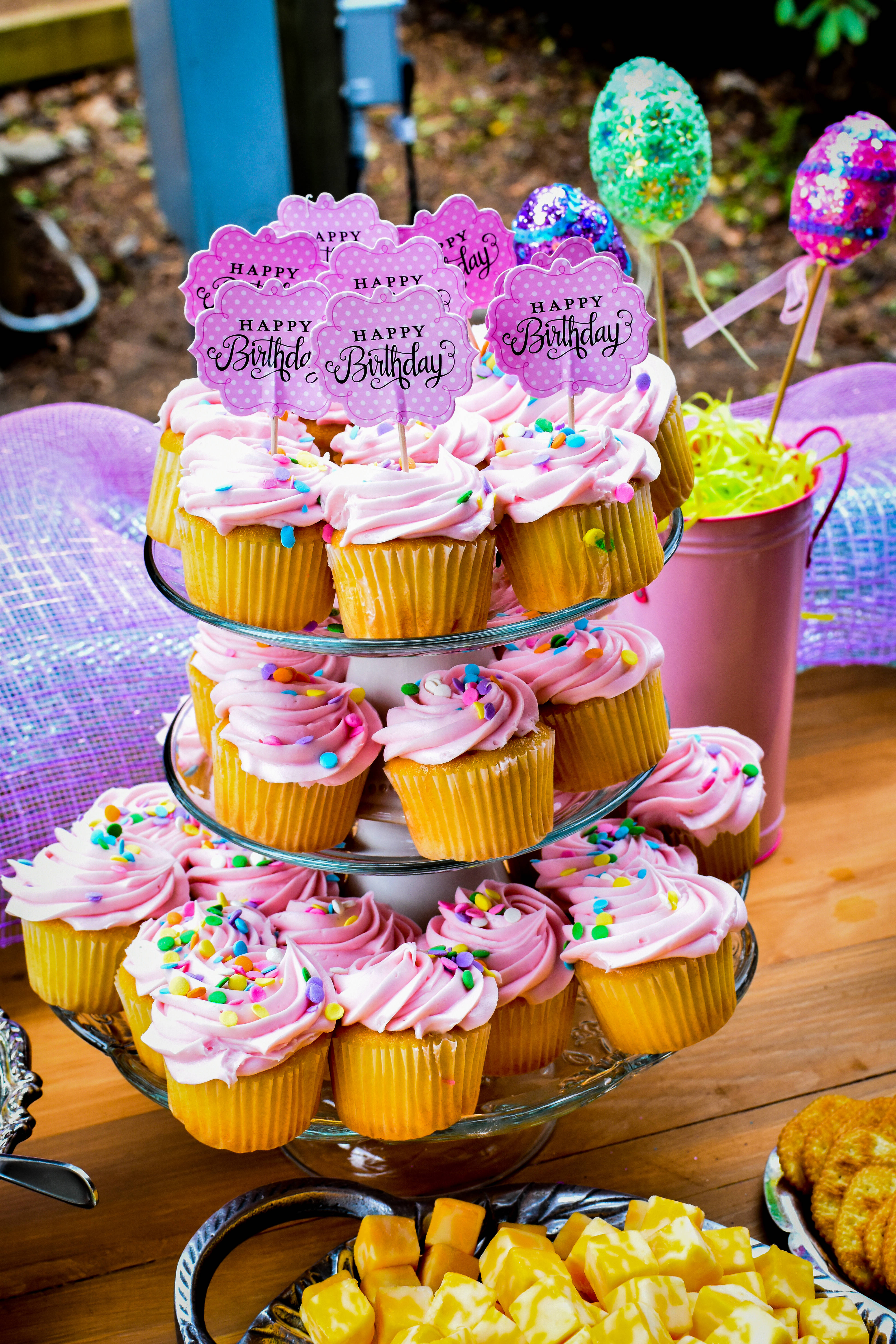 Cupcake 桌布 壁紙 — 一個充滿活力的戶外生日佈置，玻璃架上放有粉紅色糖霜杯子蛋糕，上面有彩色糖粒和「生日快樂」裝飾。