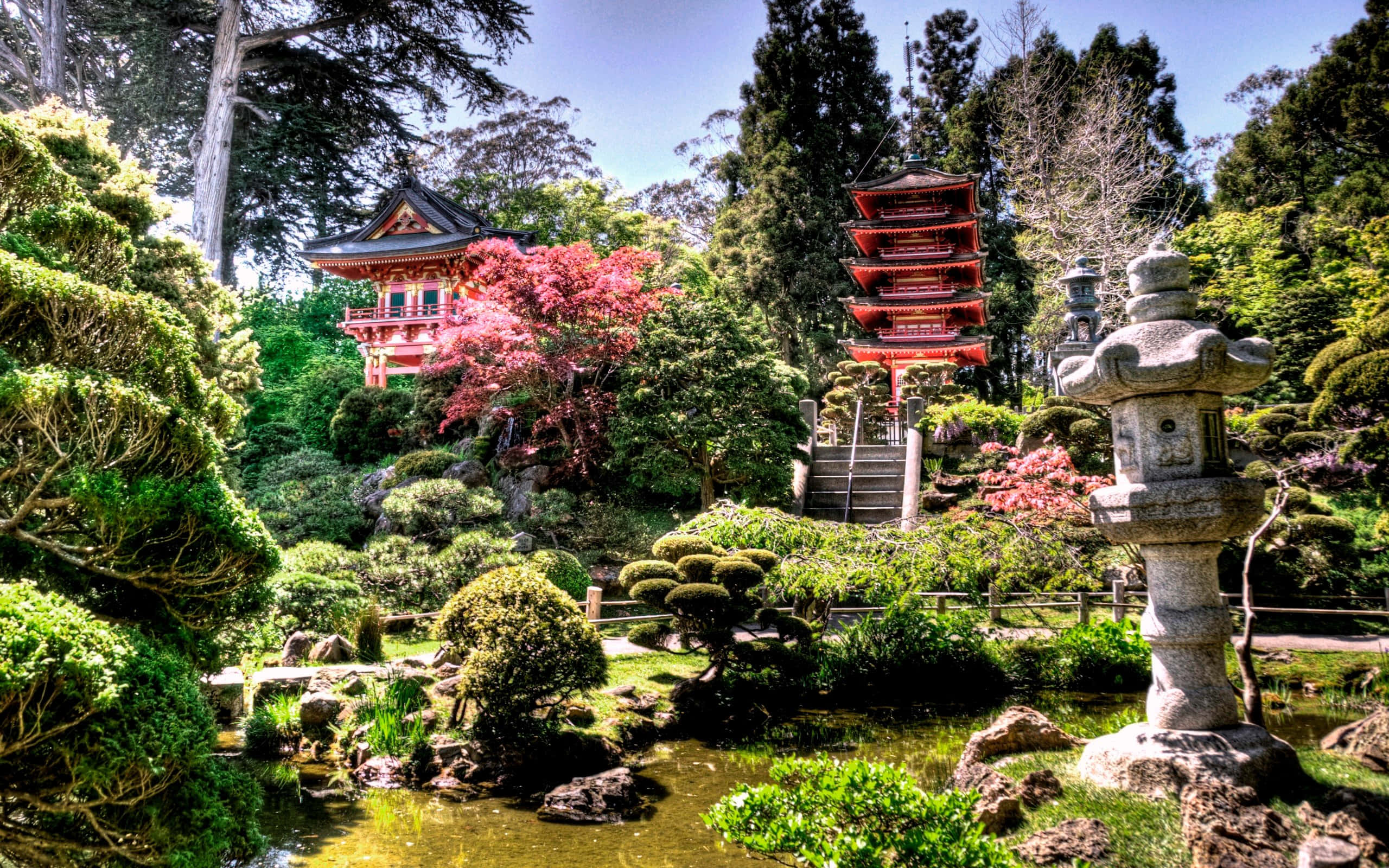 Garden 桌布 壁紙 — 一個充滿活力的日本庭園，有紅色寶塔、石燈籠和盛開的粉紅色樹木，坐落在茂密的綠色植物和寧靜的池塘之間。