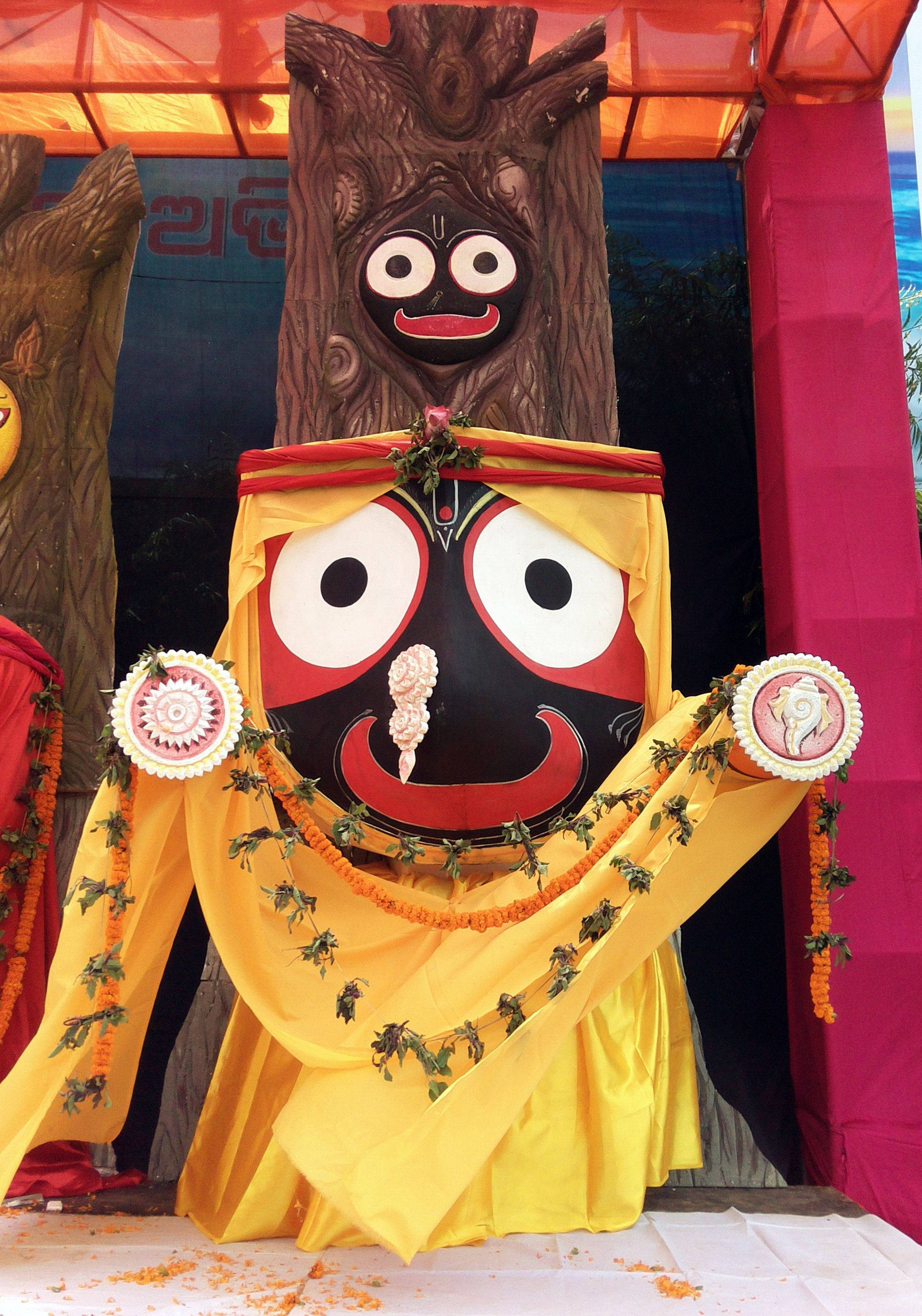 Jagannath 桌布 壁紙 — 一個充滿活力的木製Jagannath神像，披著黃布和花環，在紅色天篷下。