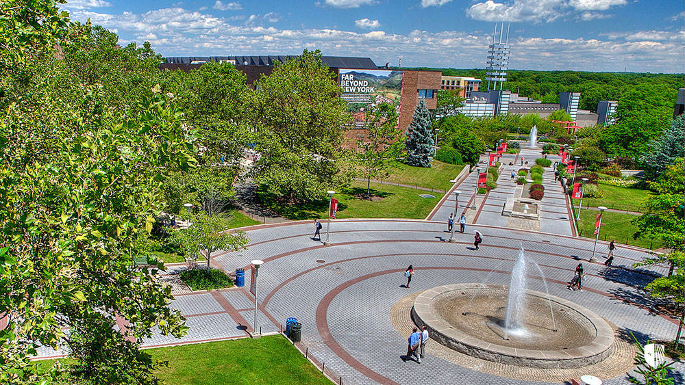 Stony Brook University 桌布 壁紙 — 一個充滿活力的校園廣場，中央噴泉周圍有學生在明亮的藍天白雲下行走。