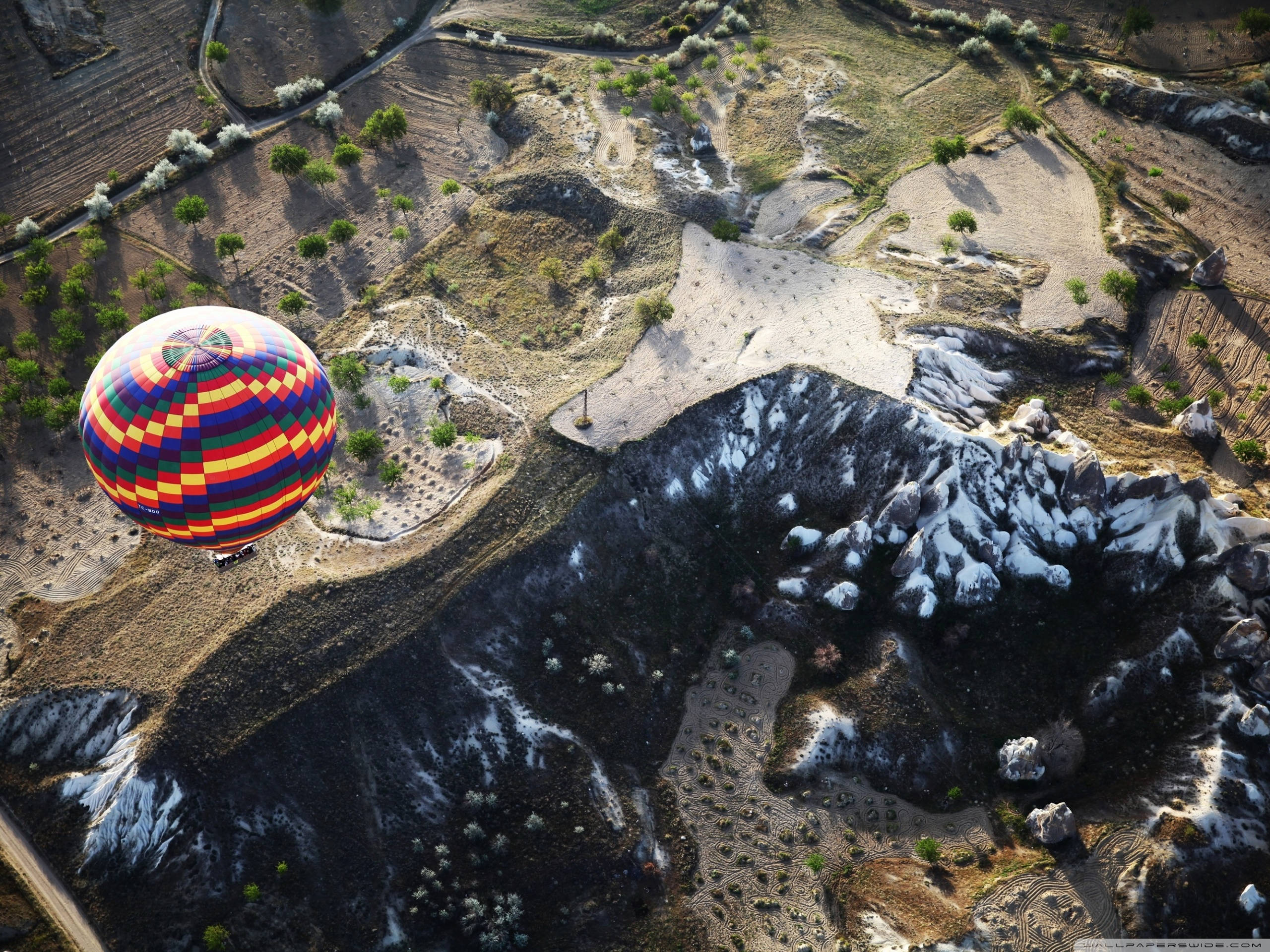Cappadocia 桌布 壁紙 — 一個充滿活力的格子熱氣球在崎嶇的火山岩層和稀疏的綠植上方，在明亮的日光下漂浮。