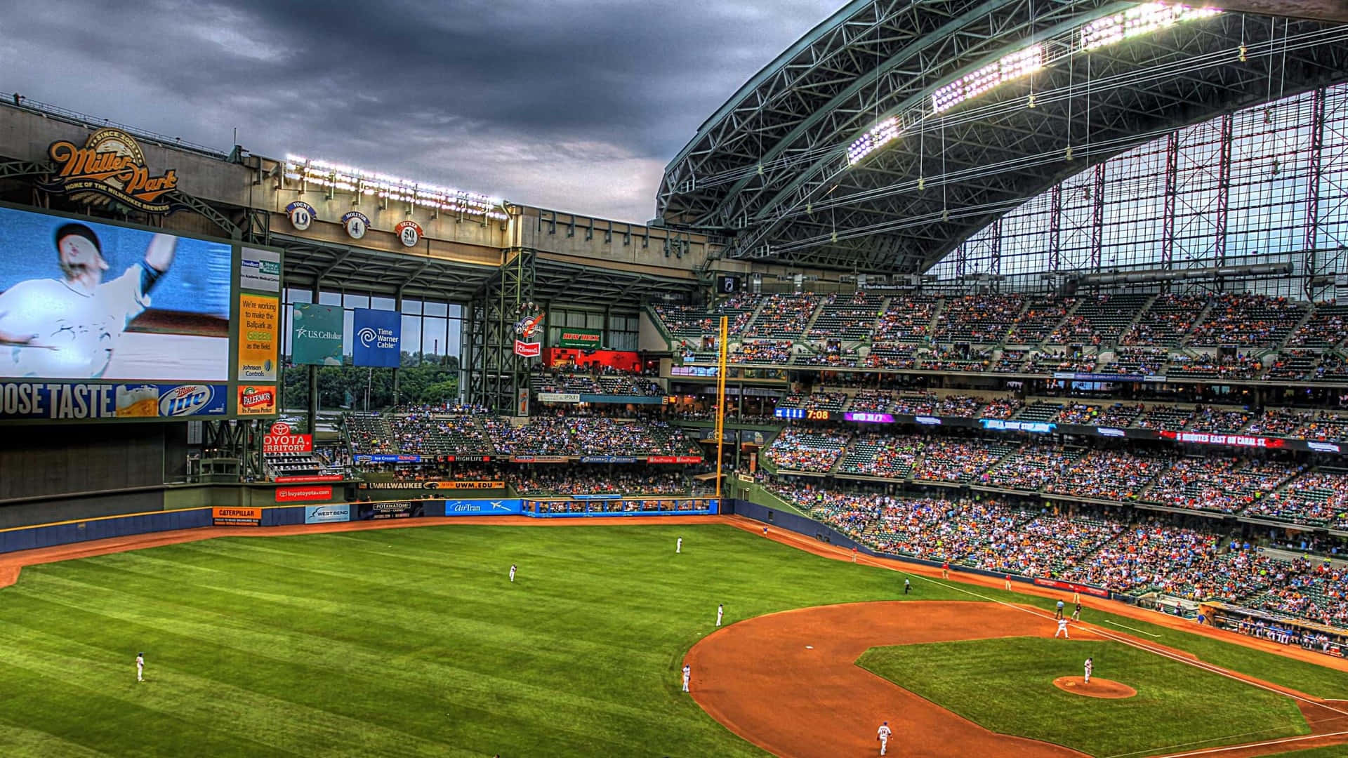 Baseball Field 桌布 壁紙 — 一個充滿活力的棒球場，在戲劇性的多雲天空下，球員在場上，看台座無虛席。