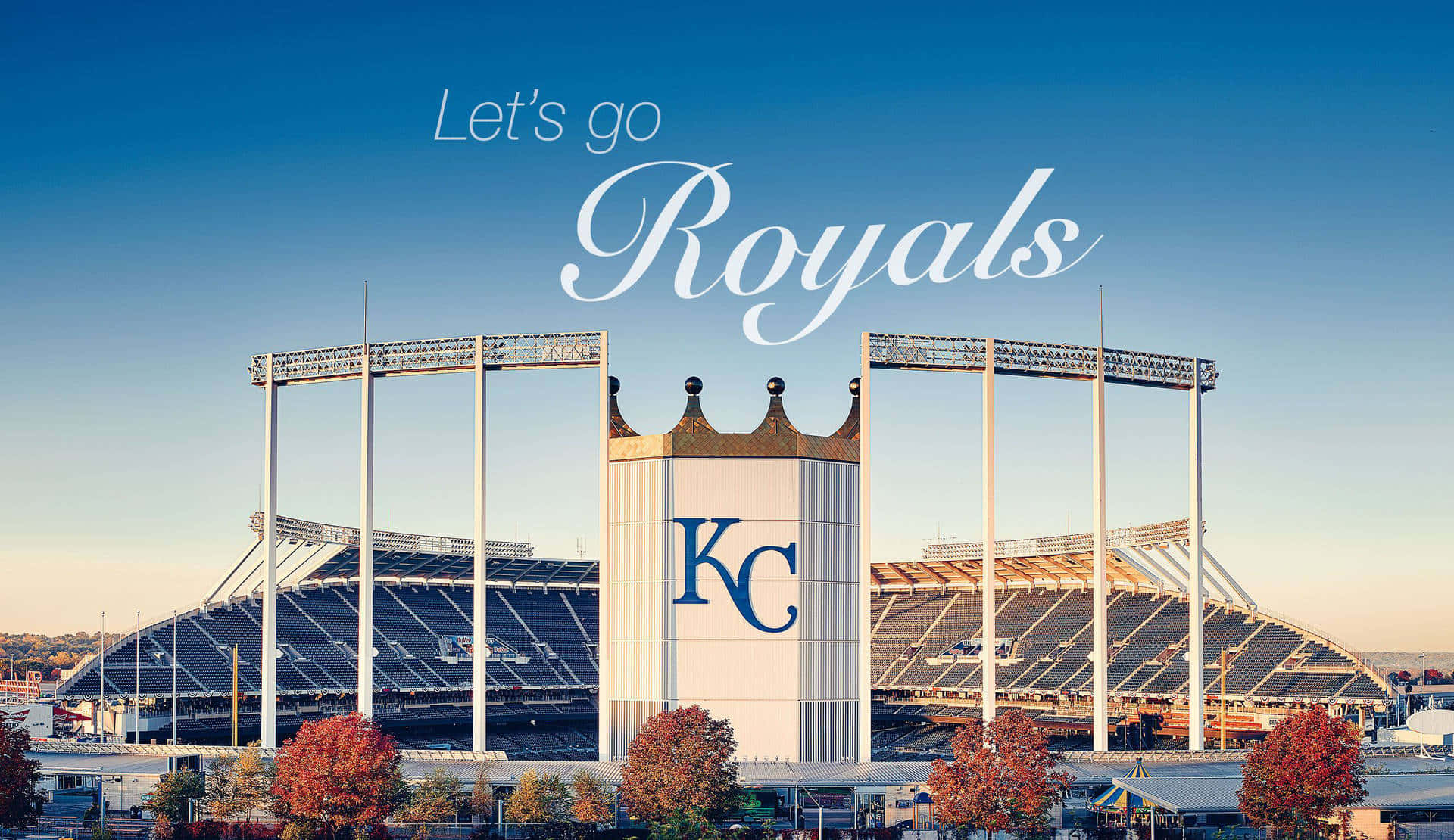 Kc Royals 桌布 壁紙 — 一個充滿活力的棒球場，帶有 'KC Royals' 品牌，在晴朗的藍天下，周圍環繞著秋天的樹木。