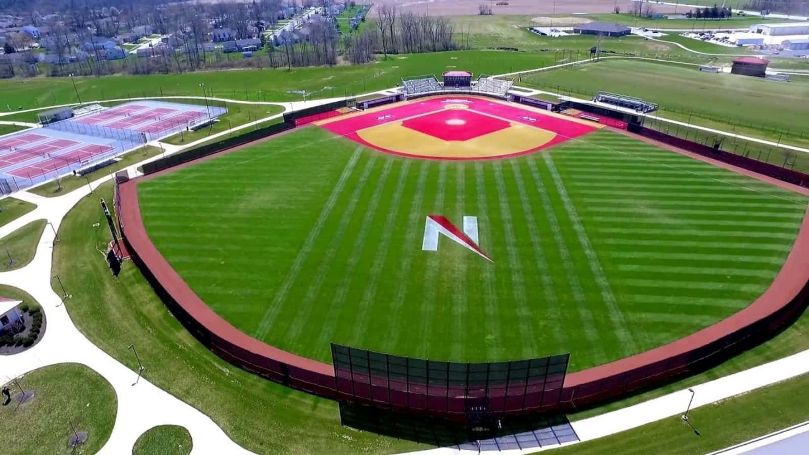 Baseball Field 桌布 壁紙 — 一個充滿活力的棒球場的鳥瞰圖，帶有紅色和黃色的鑽石形狀和綠色條紋草地，周圍環繞著網球場和郊區房屋。