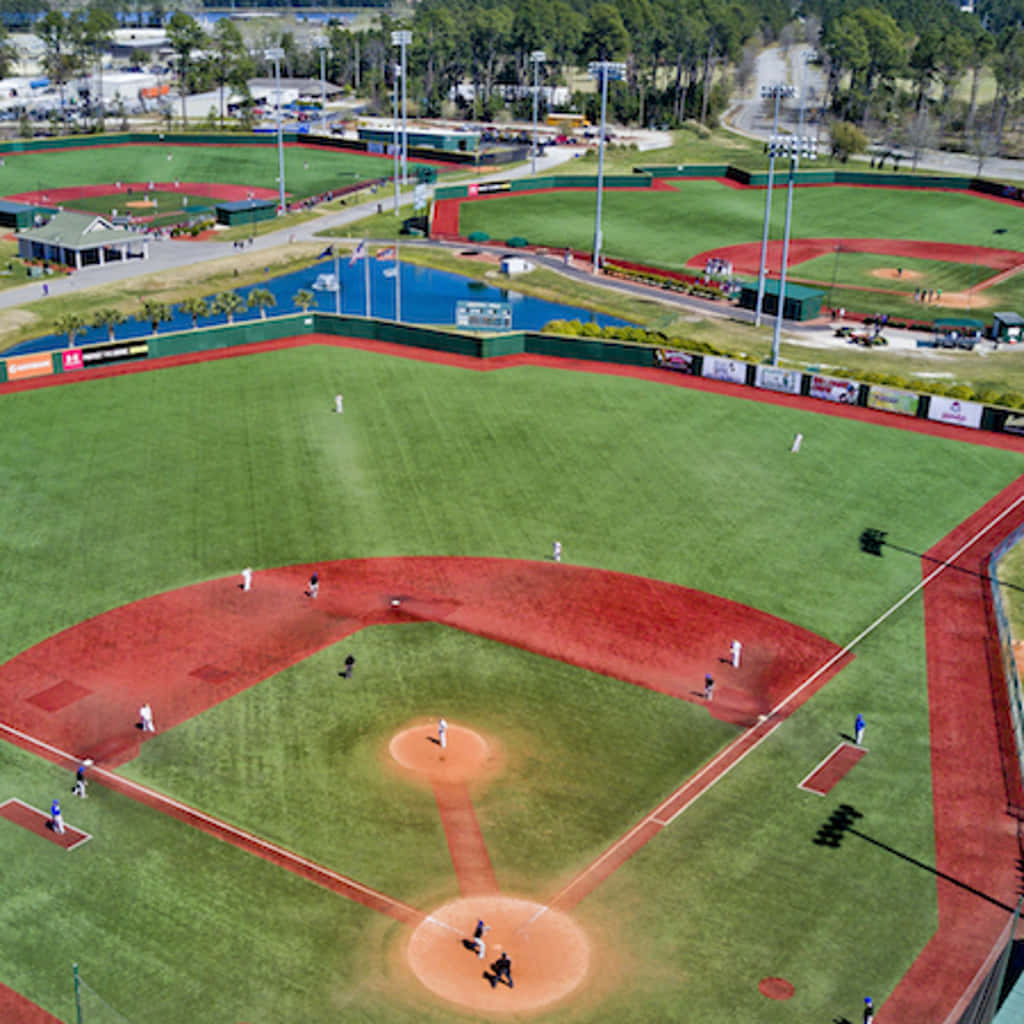 Baseball Field 桌布 壁紙 — 一個充滿活力的棒球場的鳥瞰圖，球員們正在比賽，周圍環繞著茂密的綠植和藍色水景。