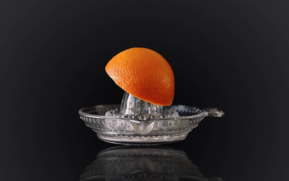 Fruits 桌布 壁紙 — 一個充滿活力的橙半邊放在玻璃榨汁機上，置於反光的黑色表面，捕捉柔和的工作室光線。