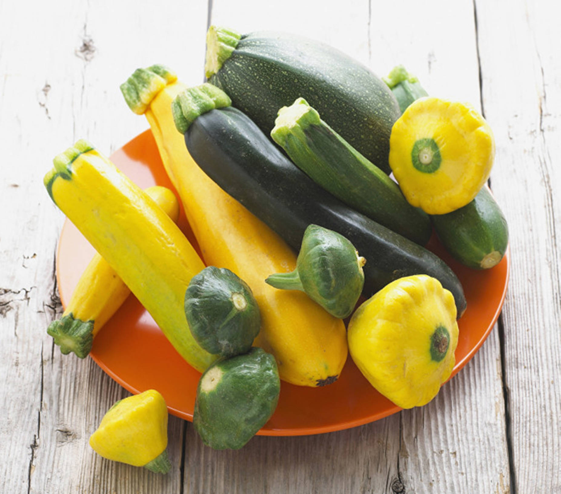 Vegetables 桌布 壁紙 — 一個充滿活力的橙色盤子，上面堆滿了黃色和綠色的櫛瓜及帕蒂潘南瓜，放在一個風化的白色木質表面上。