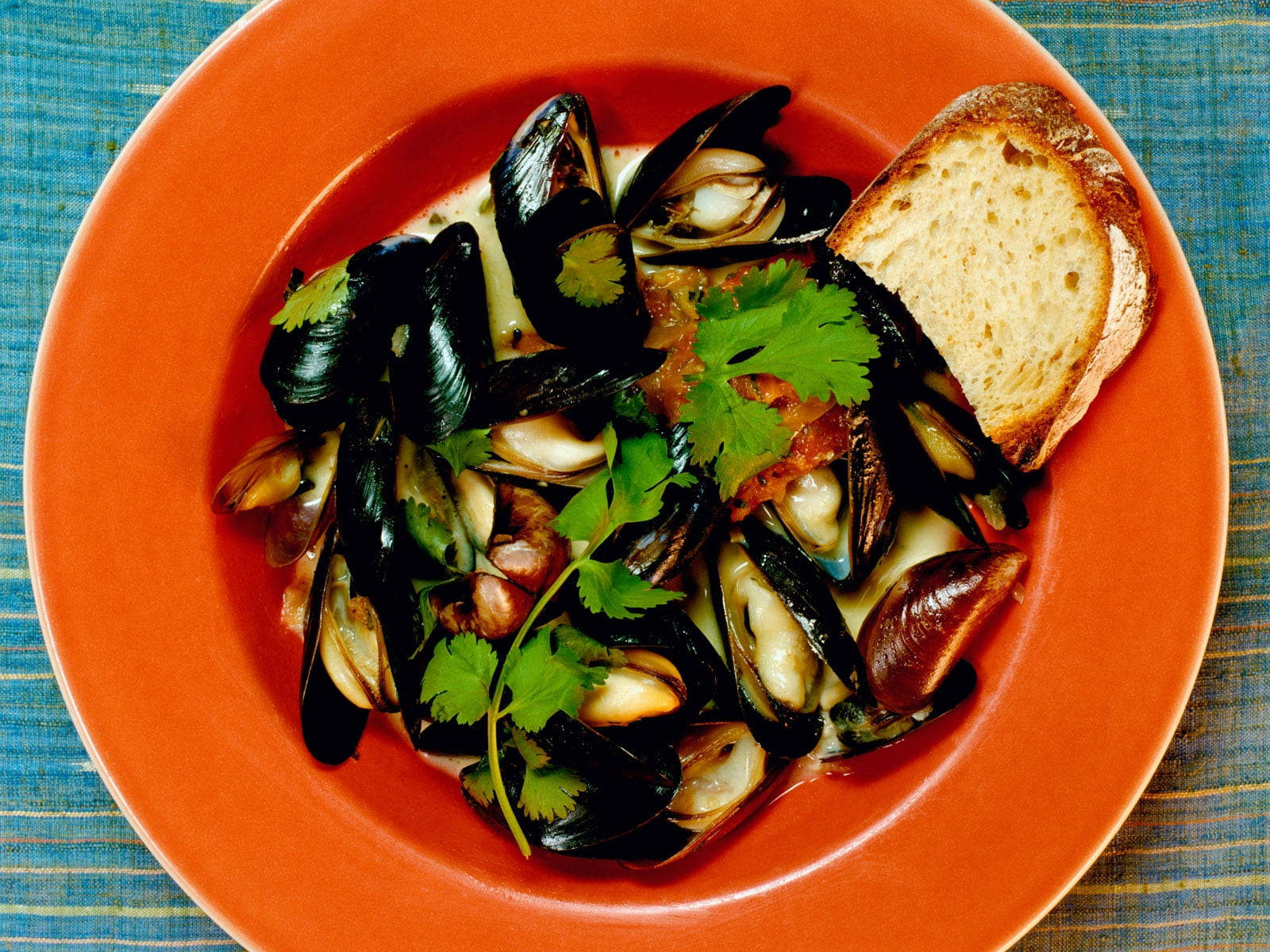 Delicious Food 桌布 壁紙 — 一個充滿活力的橙色碗盛載著以奶油醬汁蒸煮的淡菜，飾以新鮮香菜和一片脆皮麵包，放在藍色紋理桌布上。
