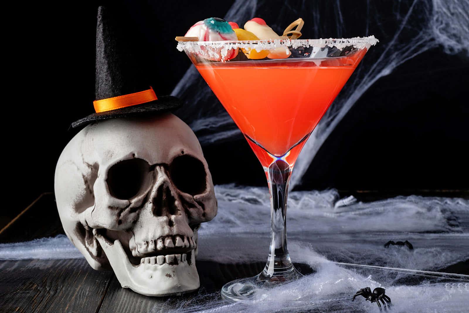 Halloween Cocktails 桌布 壁紙 — 一個充滿活力的橙色萬聖節雞尾酒放在馬丁尼杯中，旁邊是一個戴著巫婆帽的骷髏頭，放在佈滿蜘蛛網的桌子上。