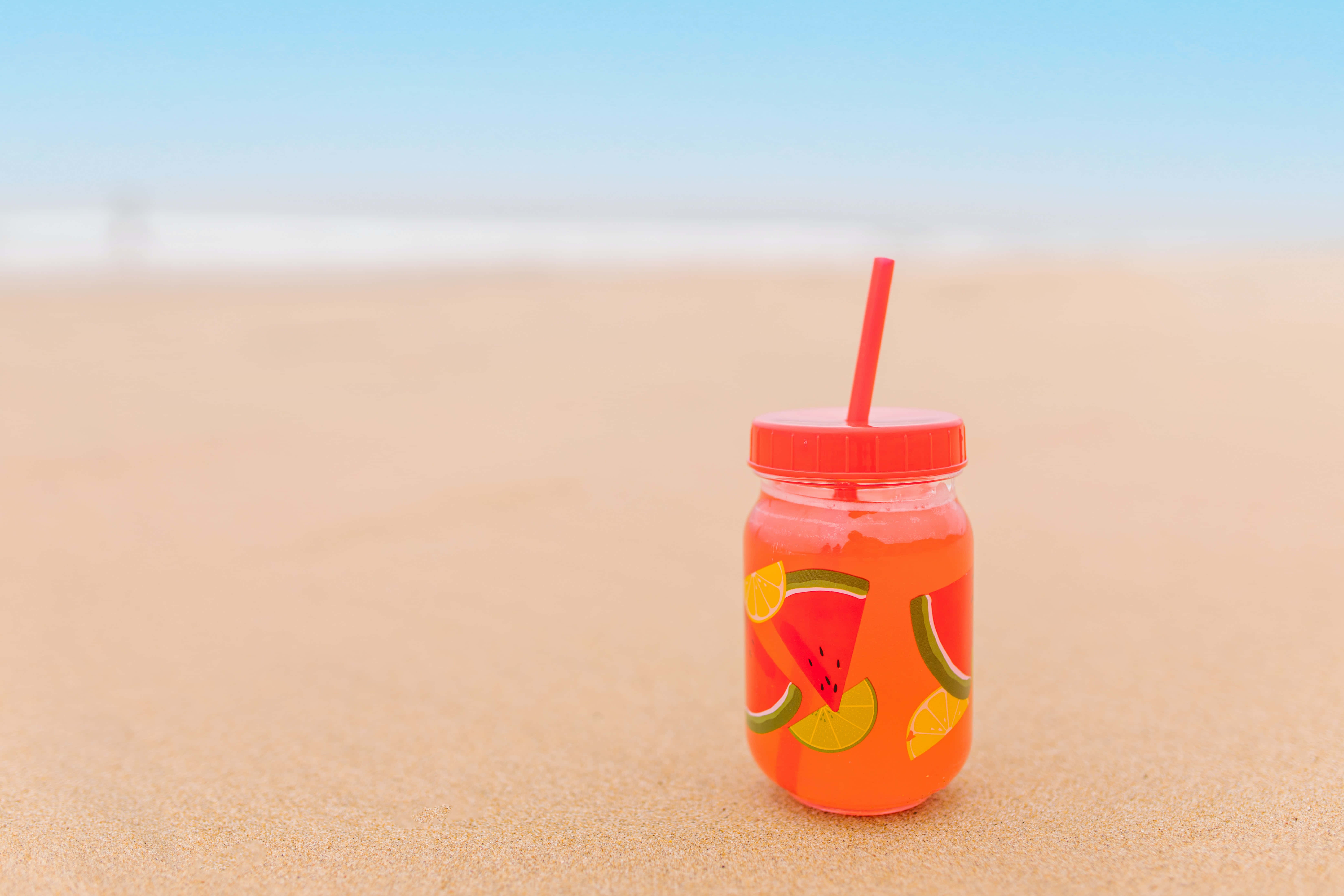 Summer Drinks 桌布 壁紙 — 一個充滿活力的橙色西瓜汁瓶，配紅色吸管，放在金色沙灘上，天空湛藍清澈。