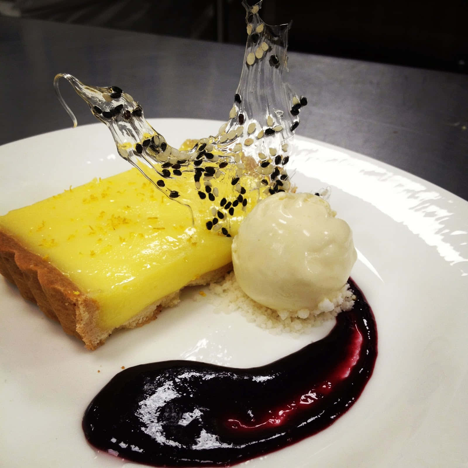 Winter Desserts 桌布 壁紙 — 一個充滿活力的檸檬塔，帶有光澤的糖霜和黑芝麻糖雕塑，旁邊是香草冰淇淋和深色莓果醬，放在白色盤子上。