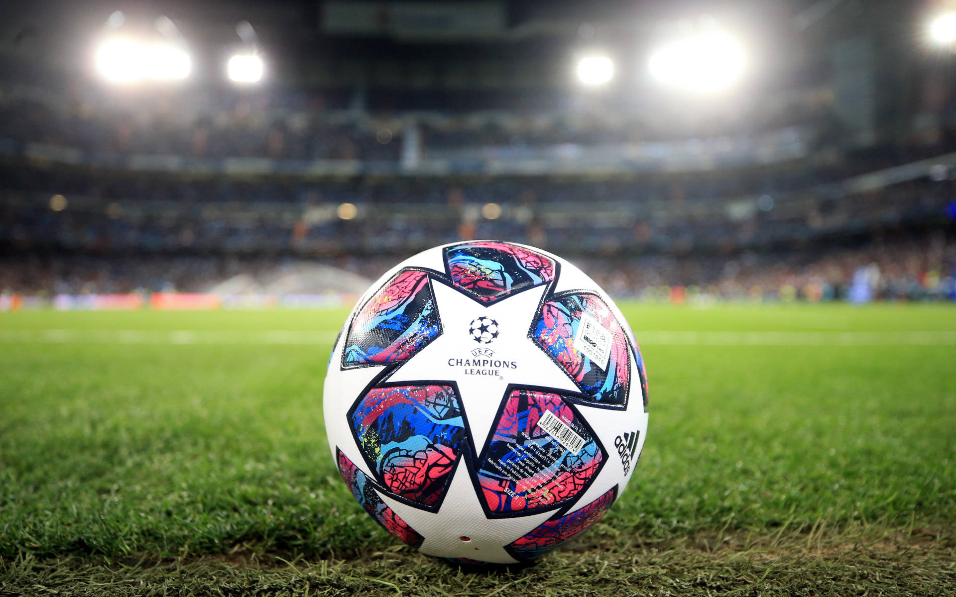 Uefa 桌布 壁紙 — 一個充滿活力的歐冠足球放在茂密的綠色球場上，在明亮的體育場燈光下，背景有模糊的觀眾。