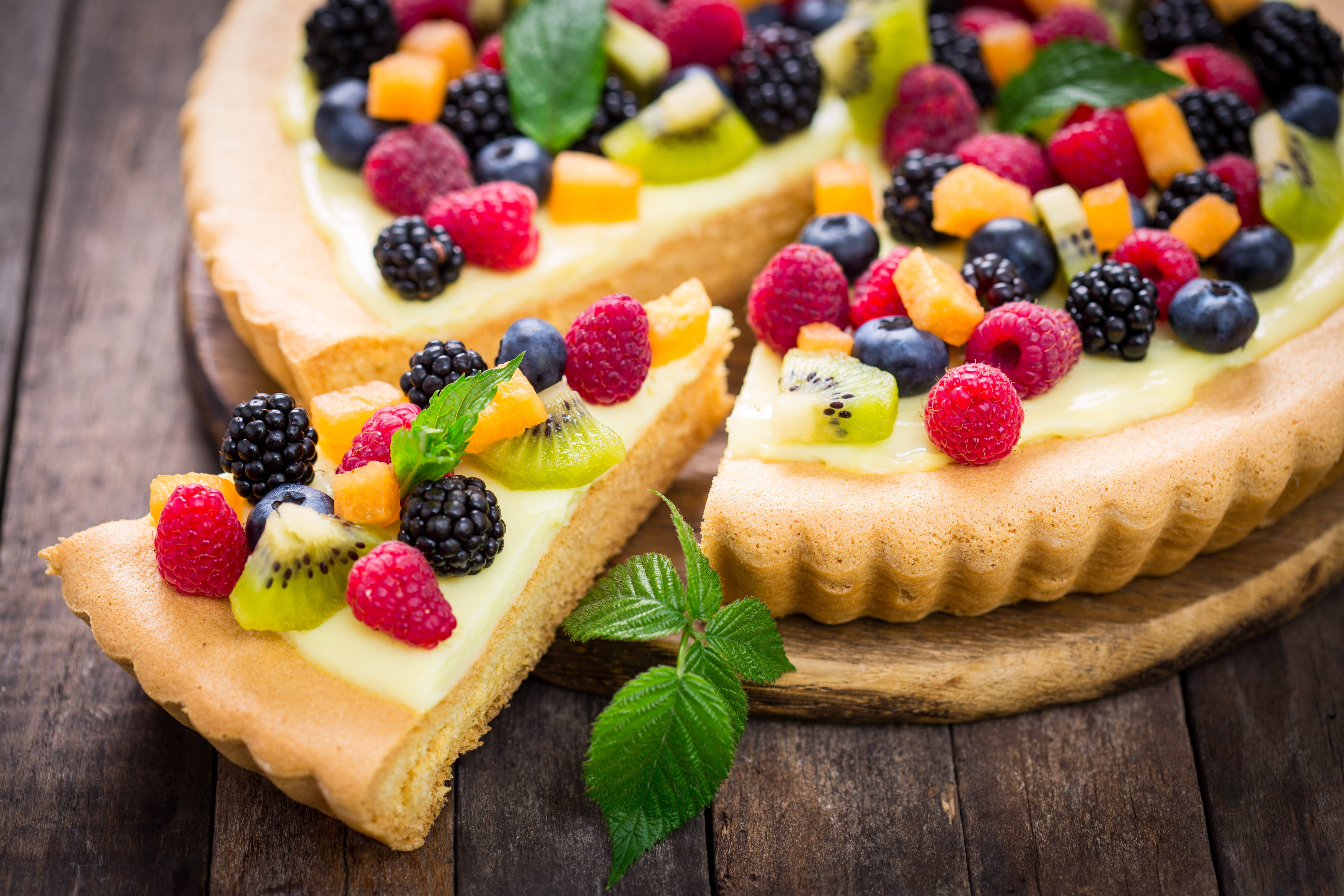 Pie 桌布 壁紙 — 一個充滿活力的水果塔，上面有覆盆子、黑莓、藍莓和奇異果片，放在一個質樸的木桌上，有一塊被切開，並用薄荷葉裝飾。