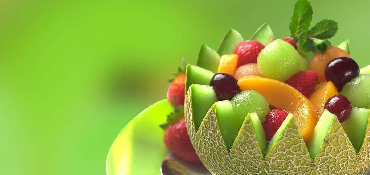 Fruits 桌布 壁紙 — 一個充滿活力的水果沙拉，放在雕刻的蜜瓜碗中，以薄荷葉點綴，襯以柔和的綠色背景。